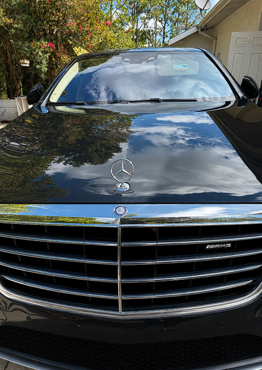 Black Mercedes-Benz car with the Mercedes logo on the hood and front grille, parked outdoors near a house with trees and flowers in the background, and a partly cloudy sky reflected on the windshield and hood.
