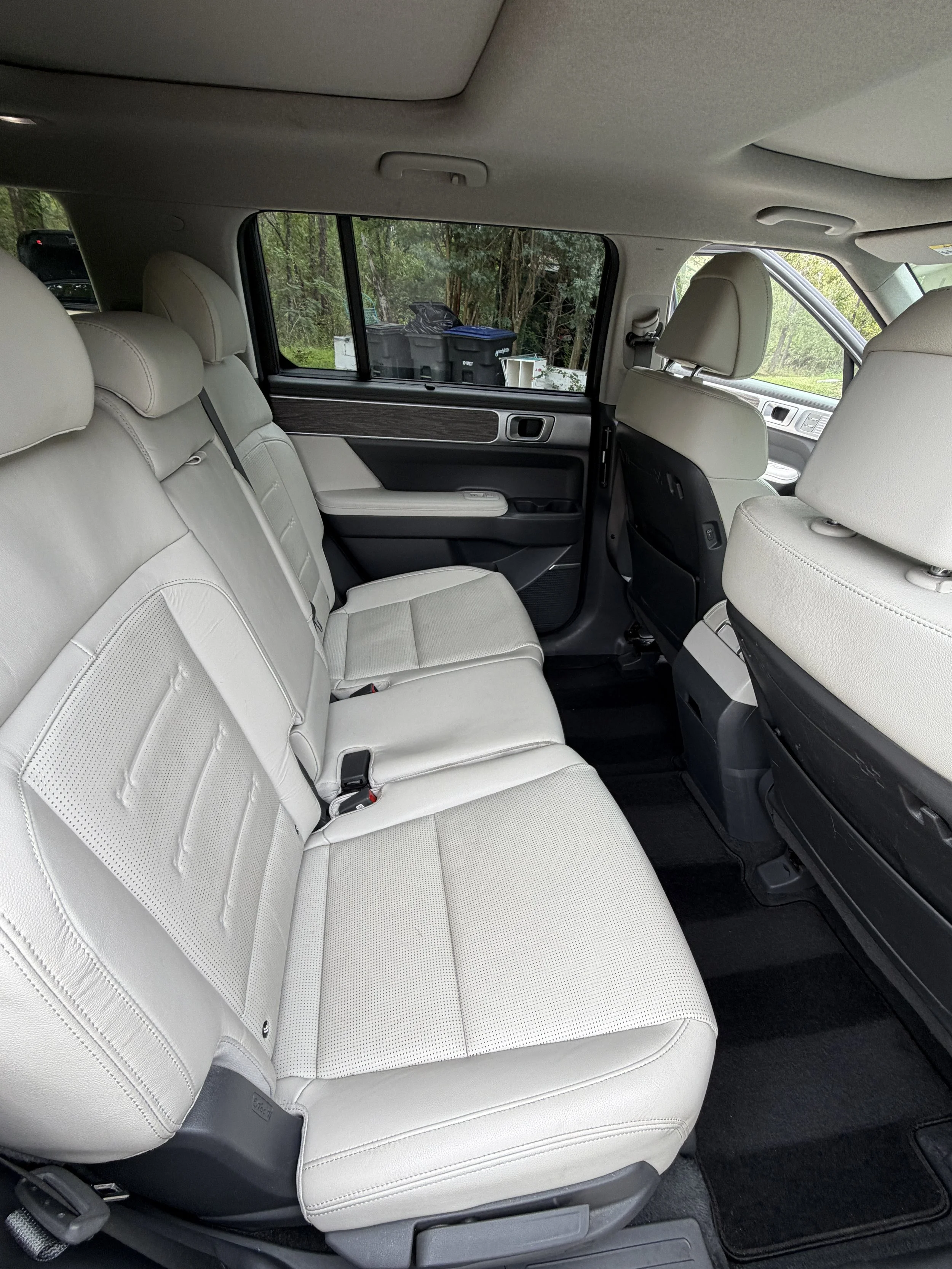 Interior of a vehicle showing beige leather seats, a black door panel with wood trim, and a window revealing outdoor trash bins surrounded by trees.