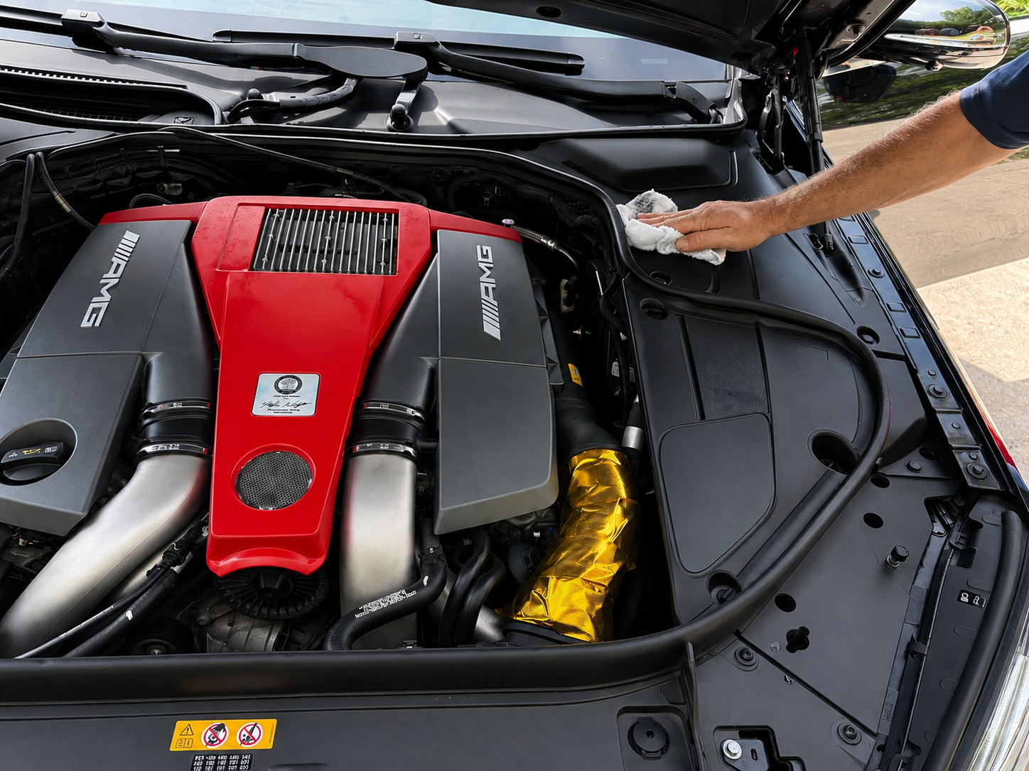 A person is cleaning the engine bay of a car with a cloth, showing a high-performance AMG engine with a red cover and silver components.