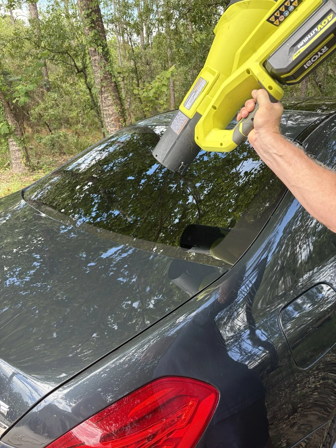 A person using a yellow leaf blower on the roof of a black car parked outdoors among trees.