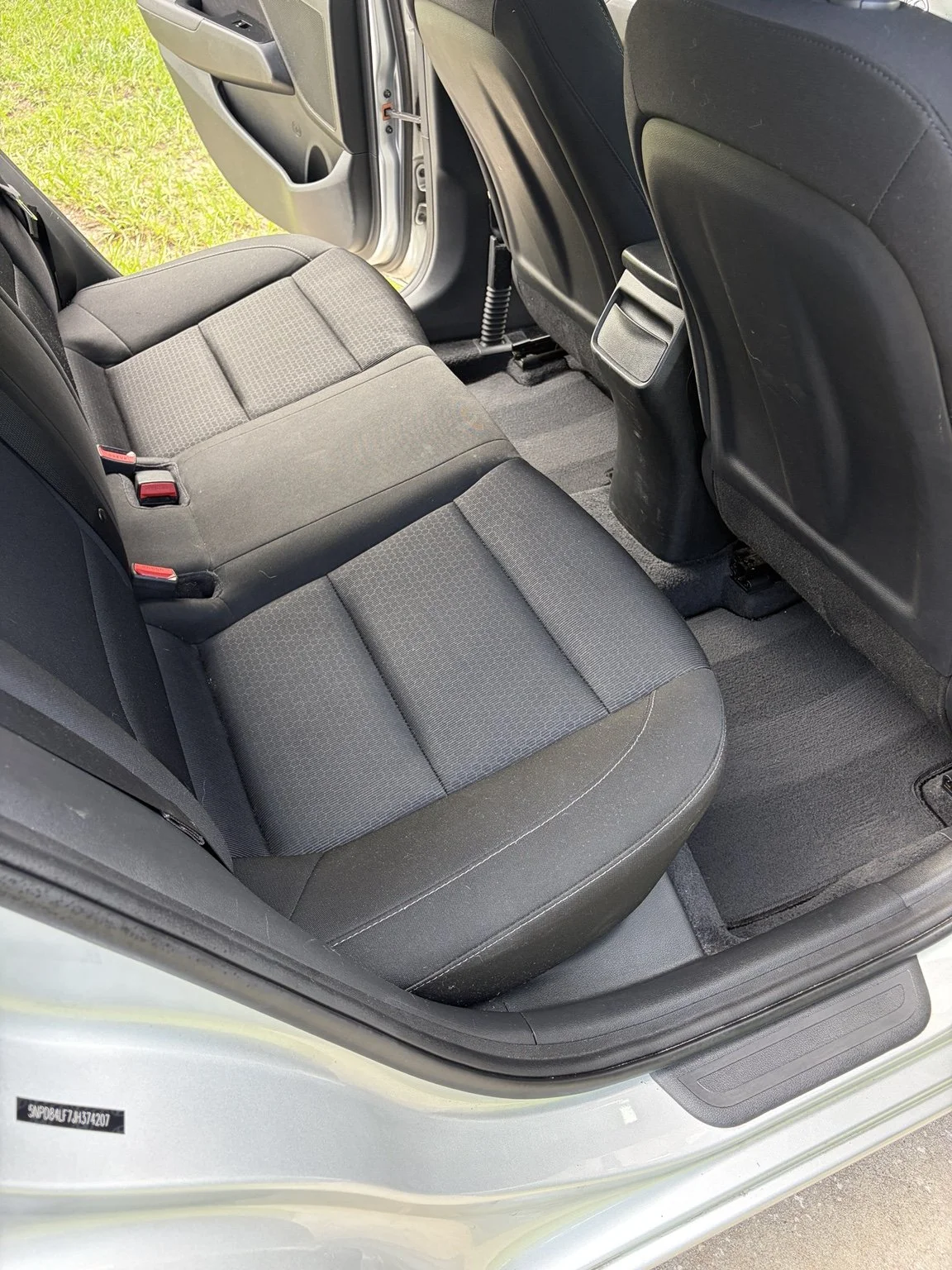 The backseat area of a car showing black fabric seats, the back of the front seats, and a view of the open rear door with green grass outside.