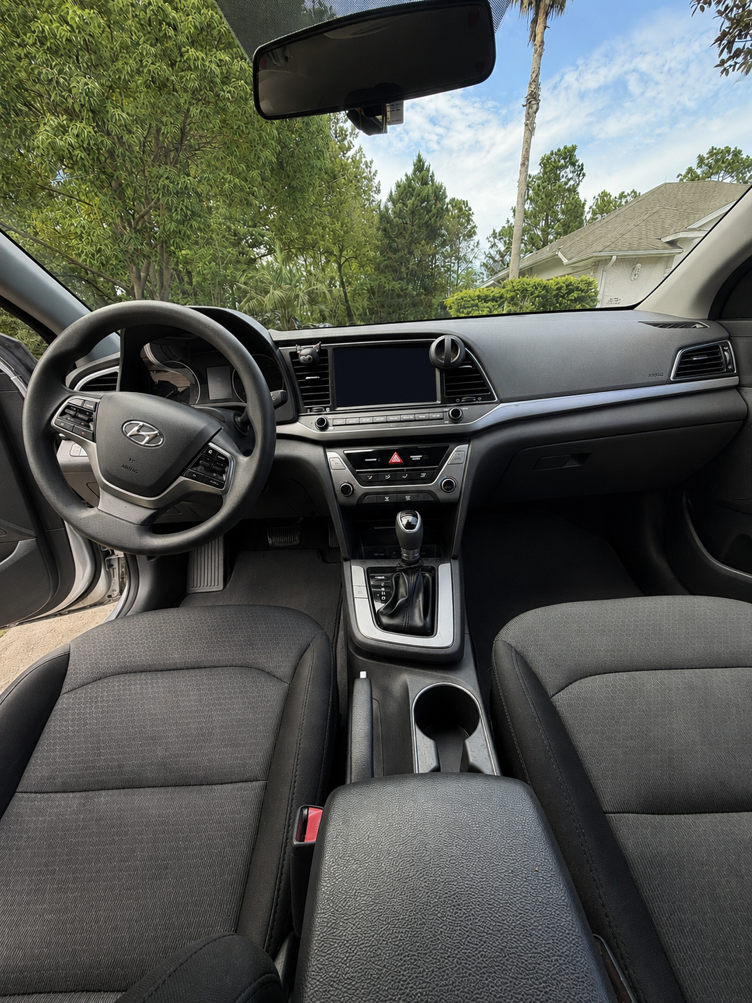 Interior of a Hyundai vehicle showing the dashboard, steering wheel, gear shift, and front seats, with greenery and a house visible through the windshield.