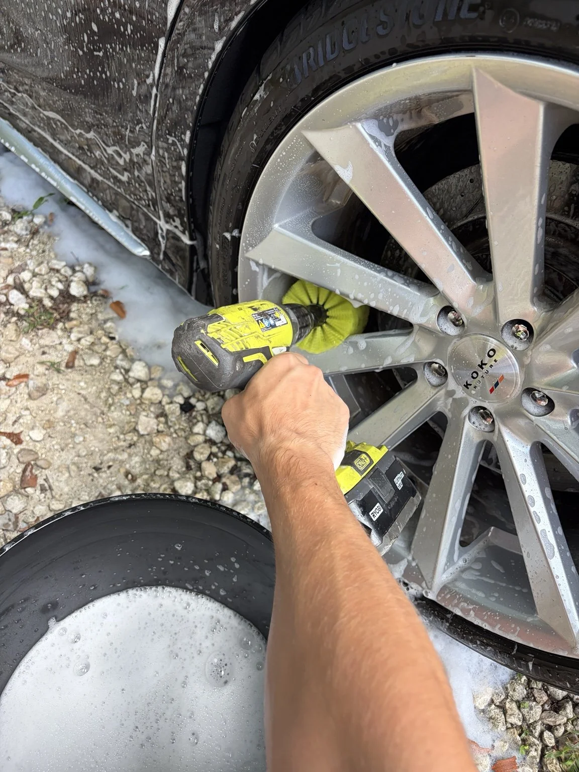 Person cleaning a silver alloy car wheel with a power drill immersed in soap foam, on a gravel surface.