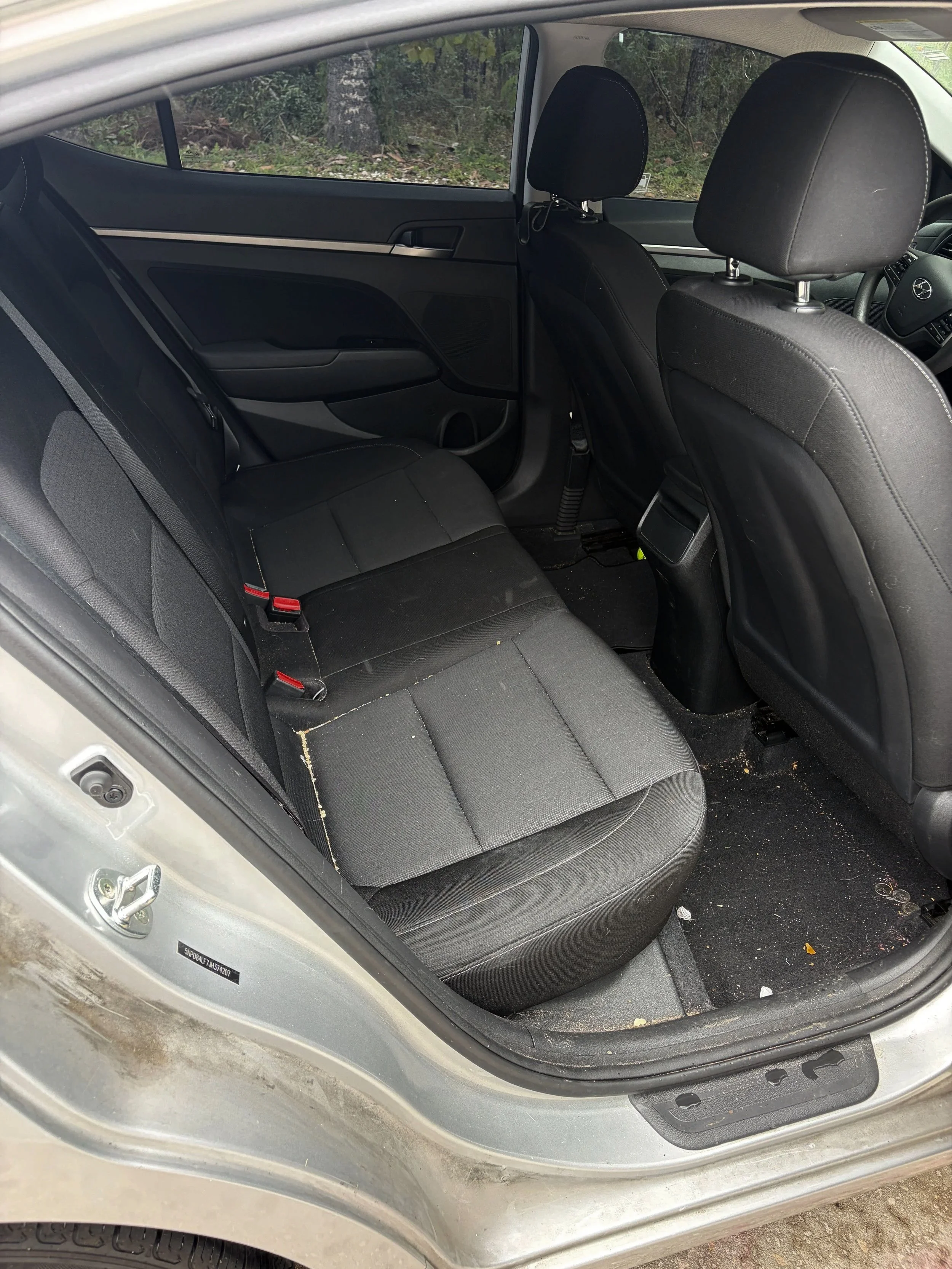 Rear passenger area before interior detailing, showing dirt and debris on the black carpet before a deep interior cleaning.