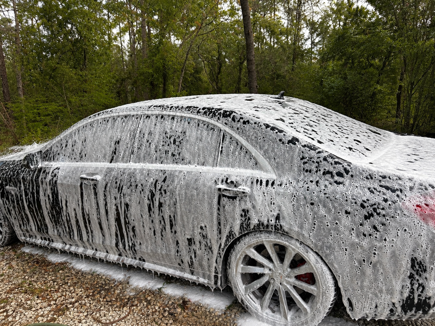 A silver car being washed with lots of soap suds covering its surface, including the windows, doors, and wheels, parked on a gravel driveway with a wooded background.