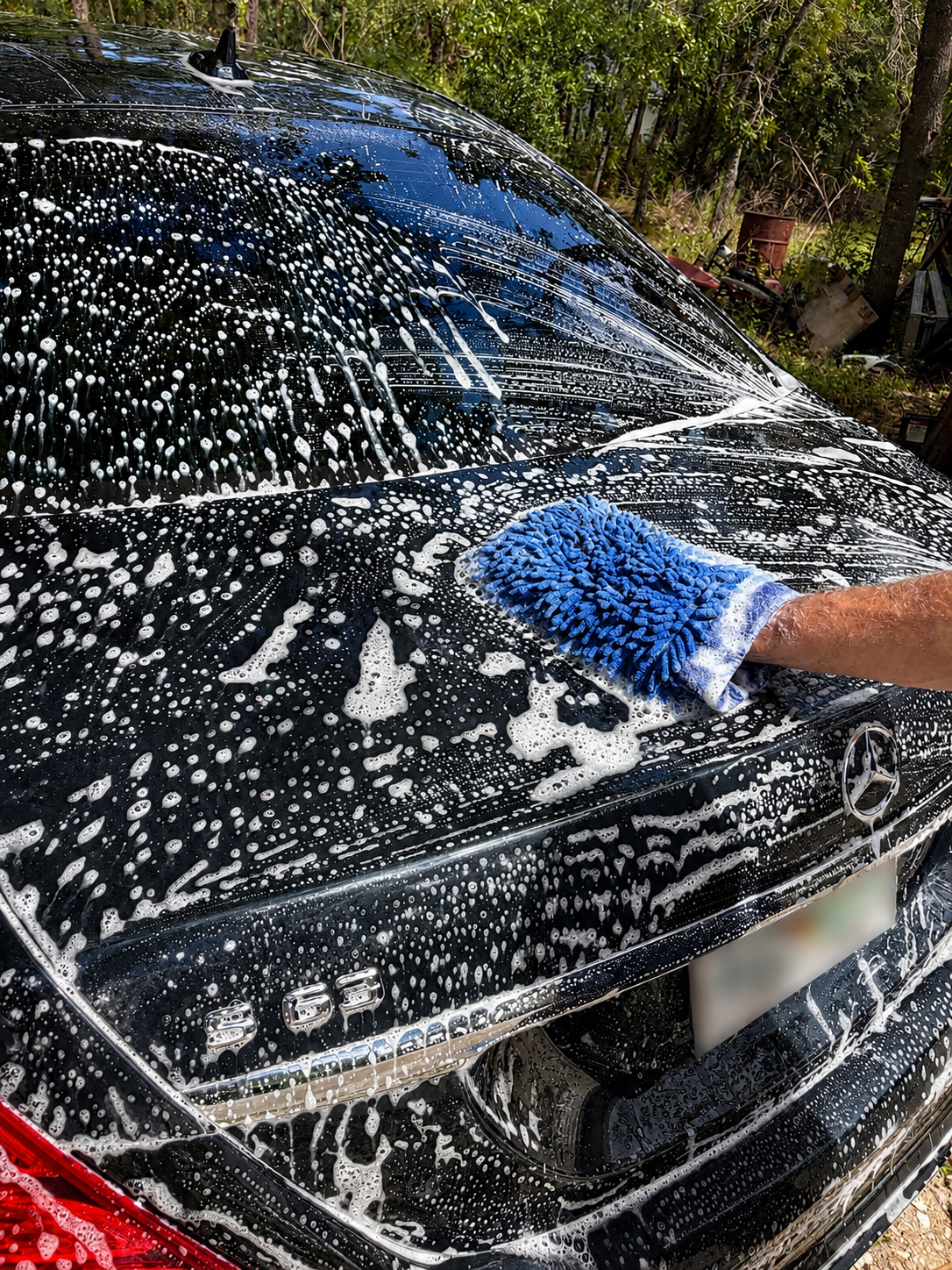 A person is washing a black Mercedes-Benz car with soapy water and a blue scrubber. The car's rear and part of the trunk are visible, with the Mercedes logo and model badge '350' on the trunk. The background shows trees and some outdoor items.