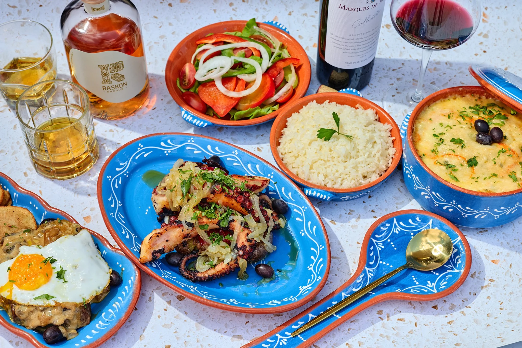 A colorful Portuguese meal on a table including rice, a salad with vegetables, a baked dish with cheese and herbs, grilled octopus with onions, a fried egg, bottles of wine and liquor, and water glasses all on a decorative table setting.