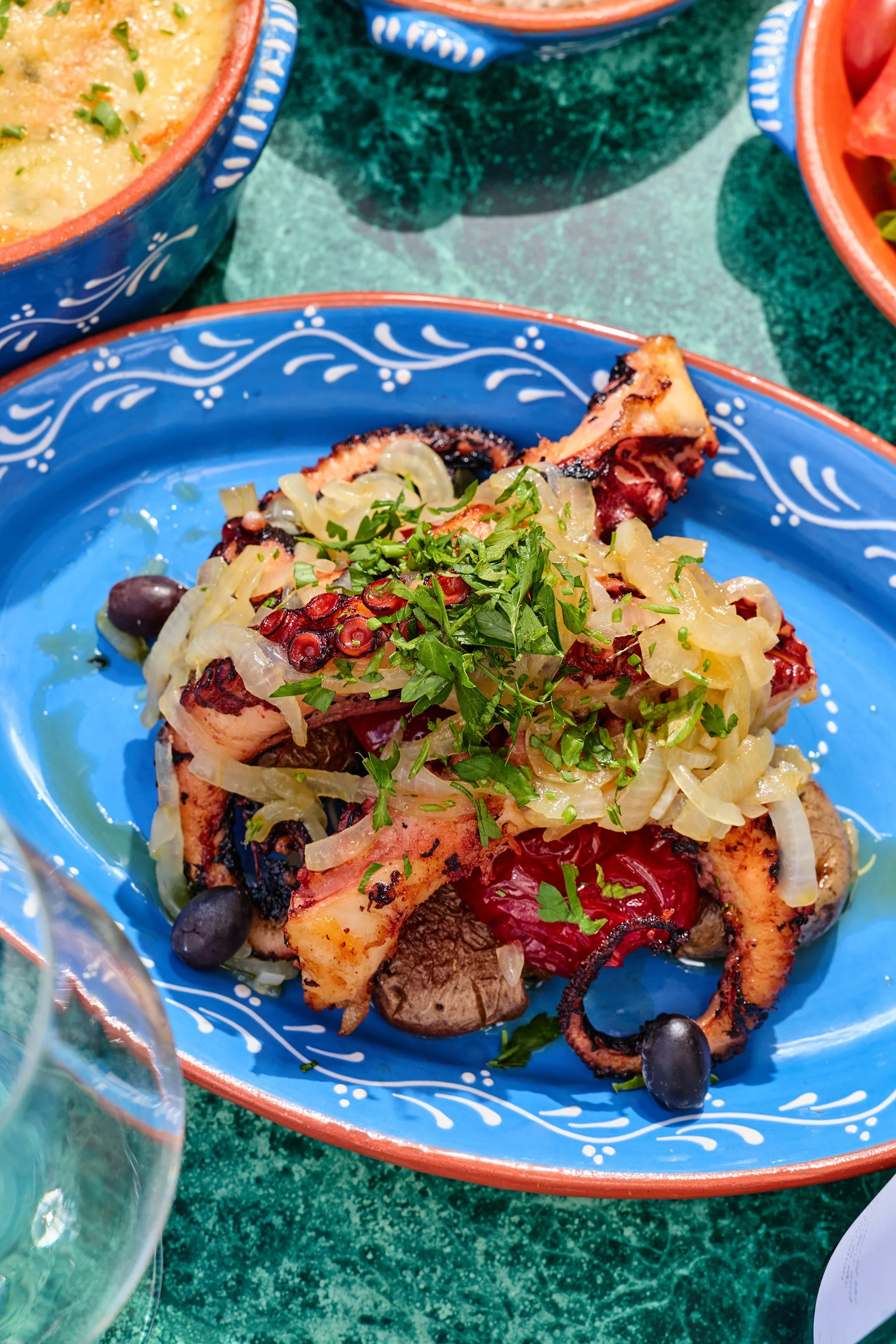 Close-up of grilled octopus with chopped onions, herbs, and berries on a blue decorative plate.