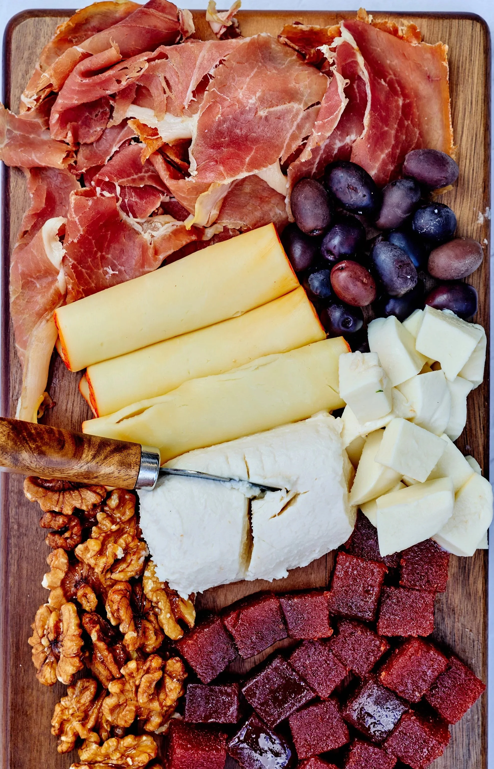 A wooden board with a variety of cheeses, nuts, fresh grapes, sliced cured meats, and snack items arranged neatly.