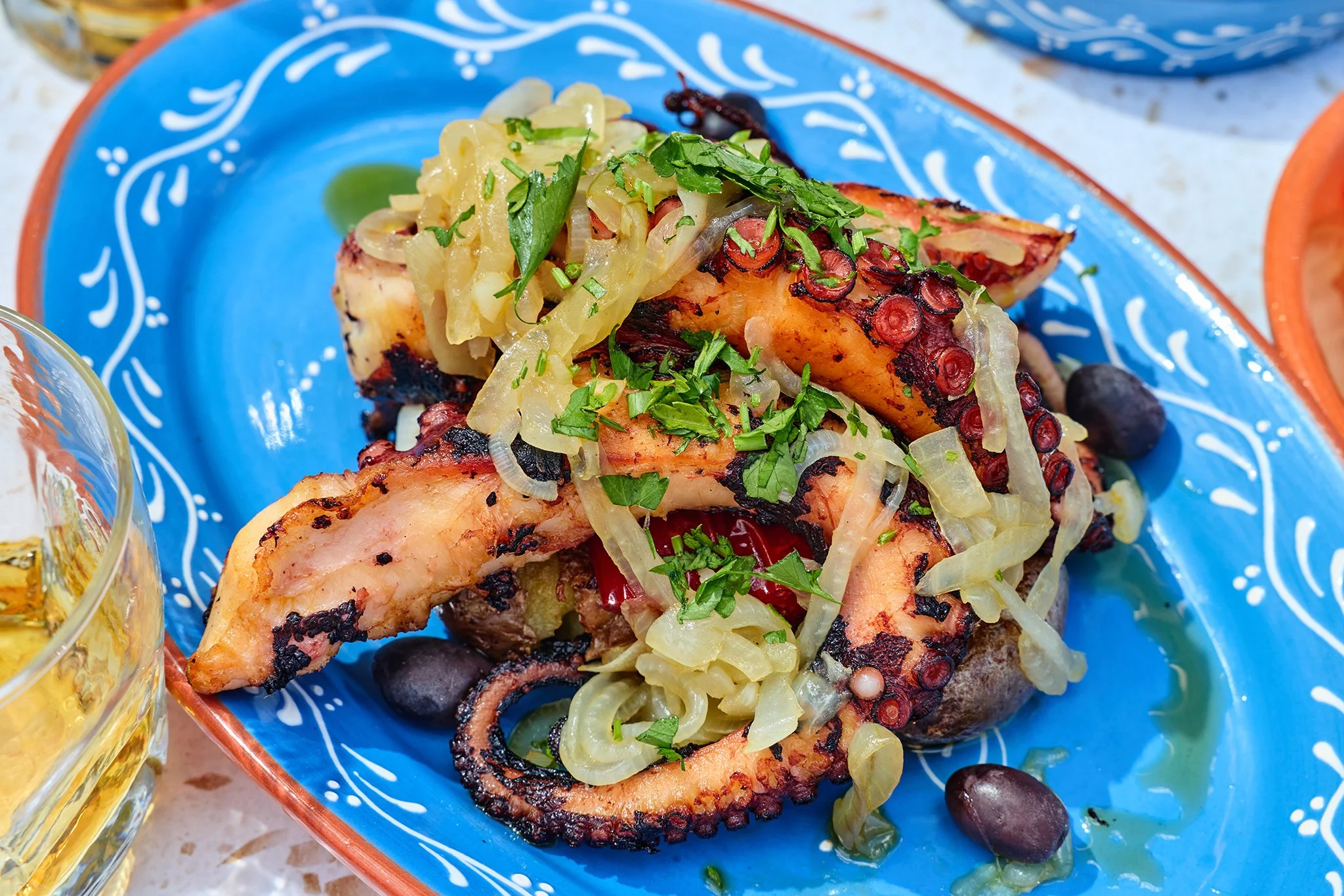 Grilled octopus tentacles topped with chopped herbs and sliced onions on a blue and white ceramic plate with a side of olives.