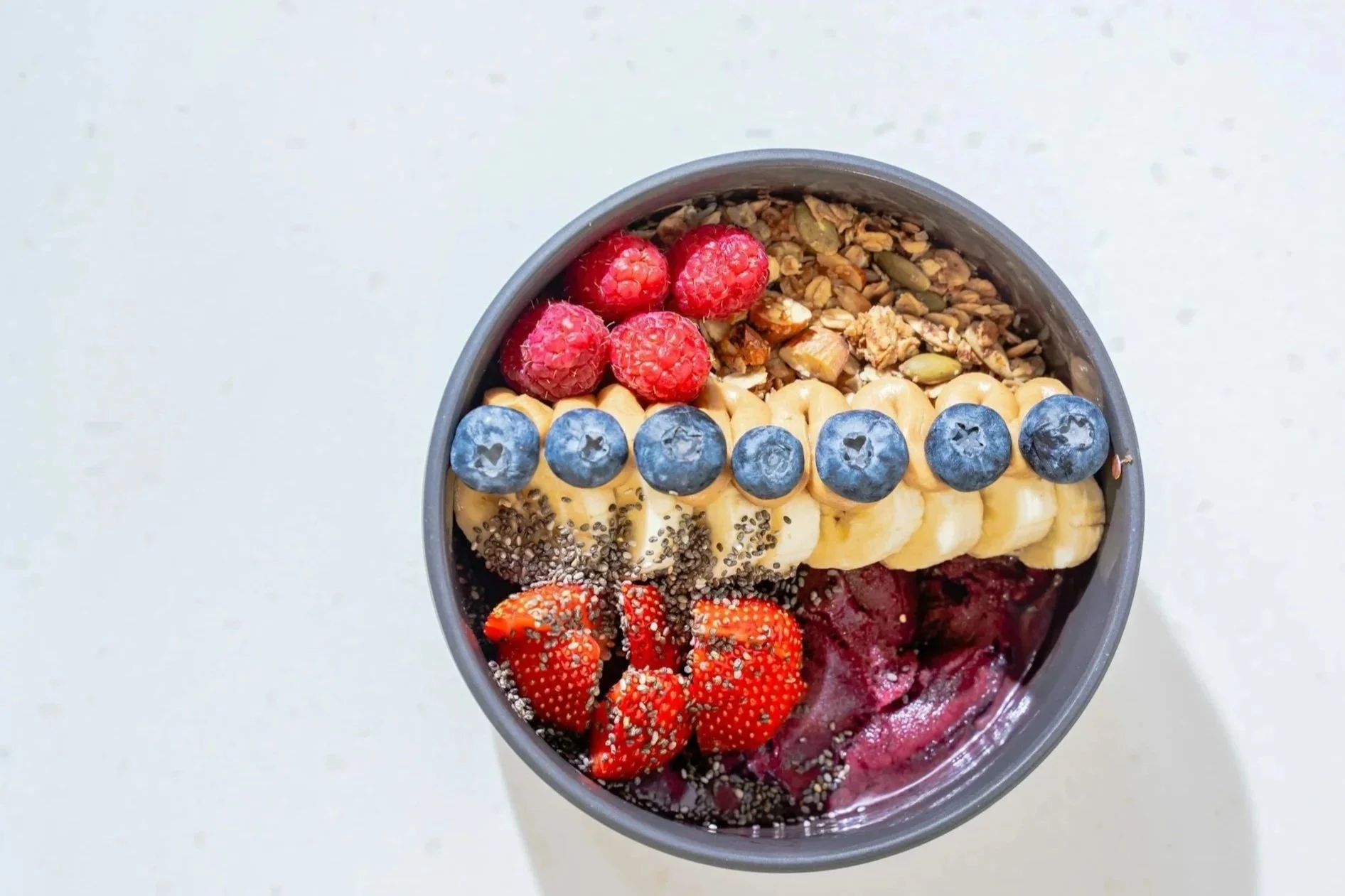 Overhead view of a acai bowl topped with fresh berries, banana slices, granola, seeds, and chia seeds, with a white background.
