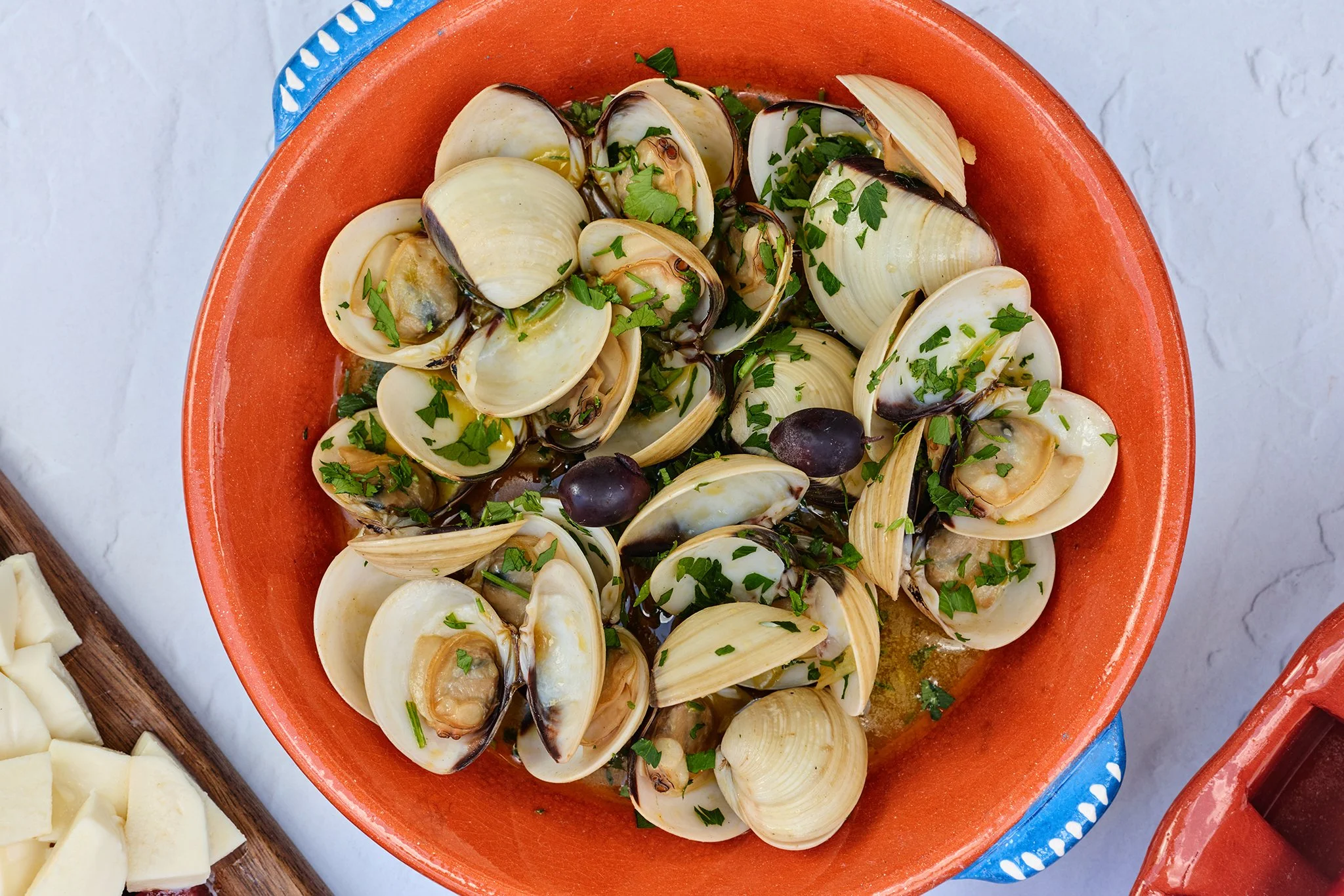 A bowl of cooked clams garnished with chopped parsley, with slices of cheese and a cutting board nearby.