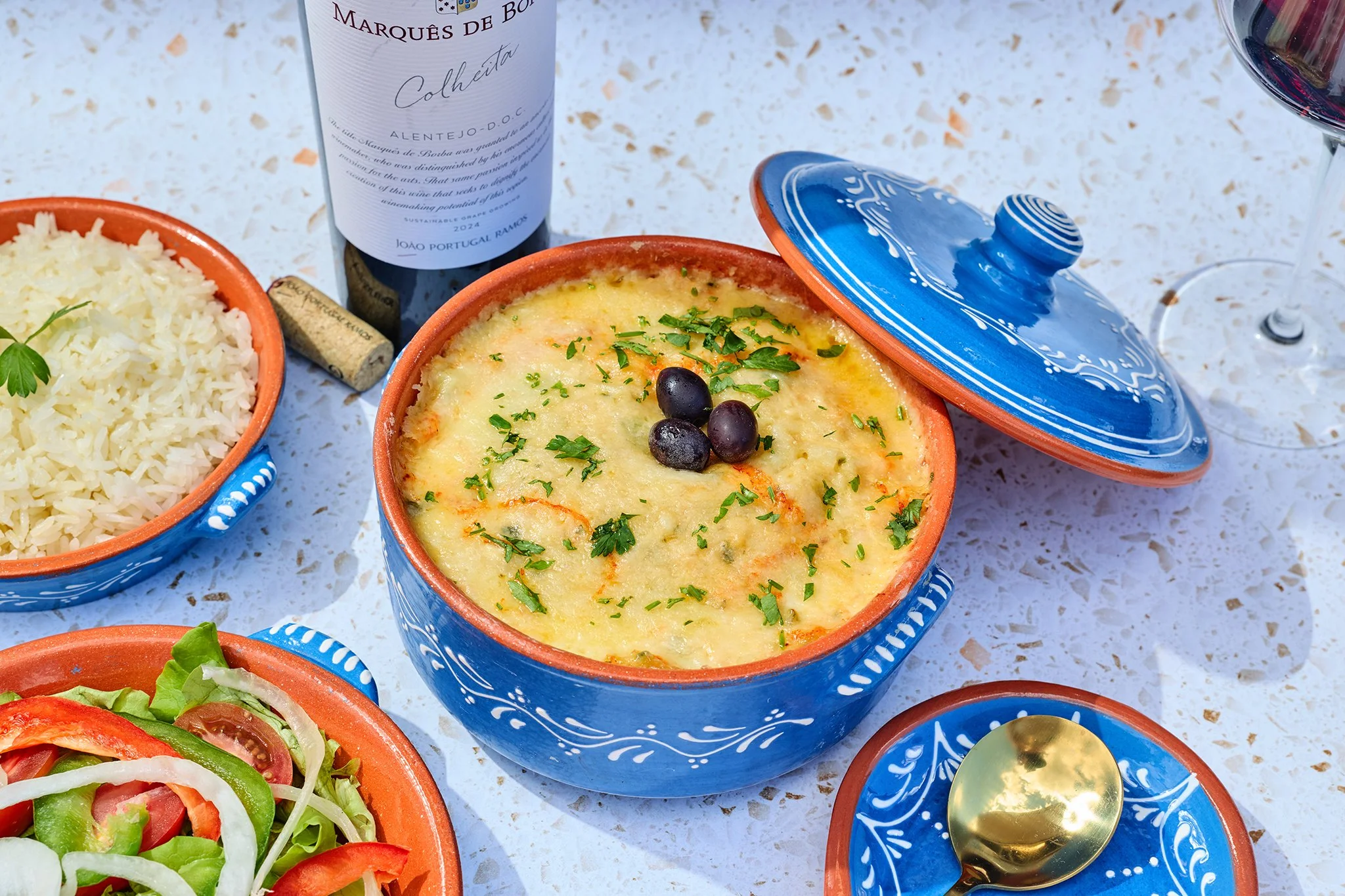 Traditional Portuguese casserole dish in a blue ceramic pot with a partially removed lid, garnished with chopped herbs and black olives, next to a bowl of rice, with plates of fresh salad, wine, and a bottle of wine in the background.