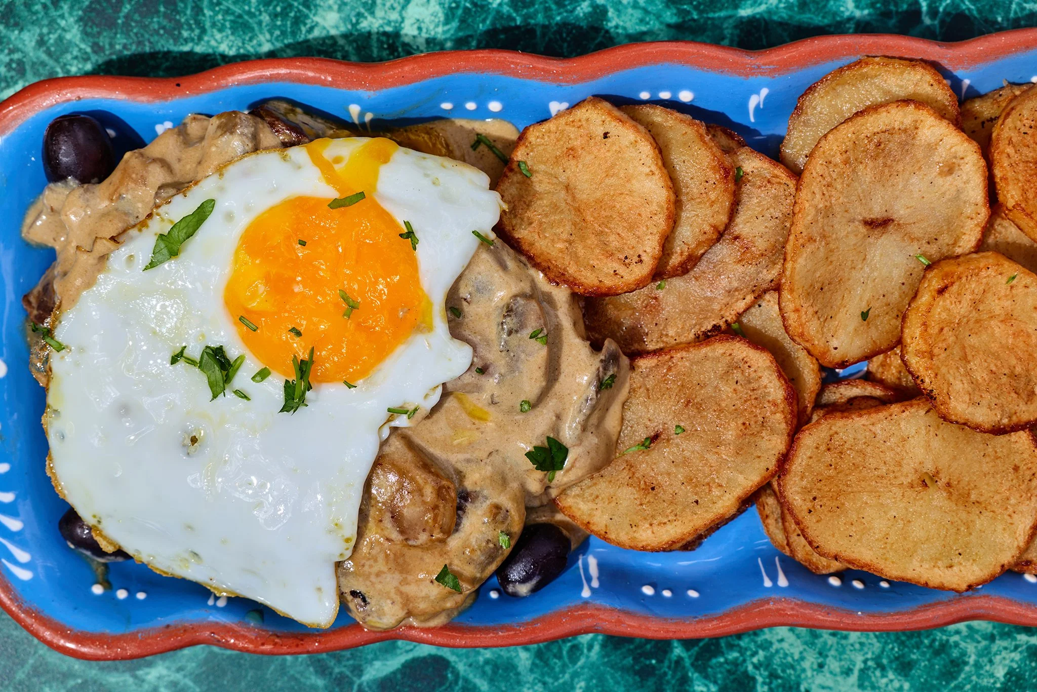 A colorful bowl with a fried egg, potatoes, and a creamy dish with mushrooms, garnished with chopped herbs.