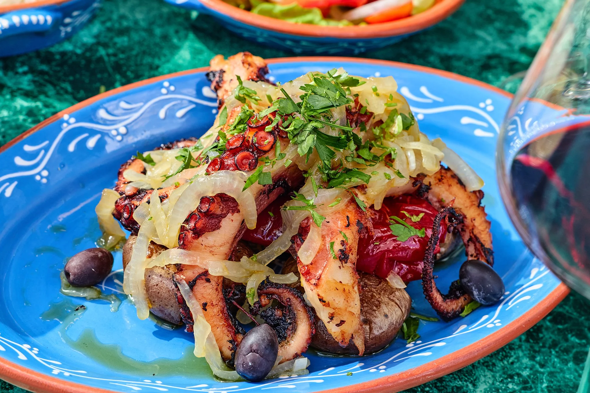 Grilled octopus topped with chopped herbs, grilled octopus, sautéed onions, and black olives served on a colorful plate.