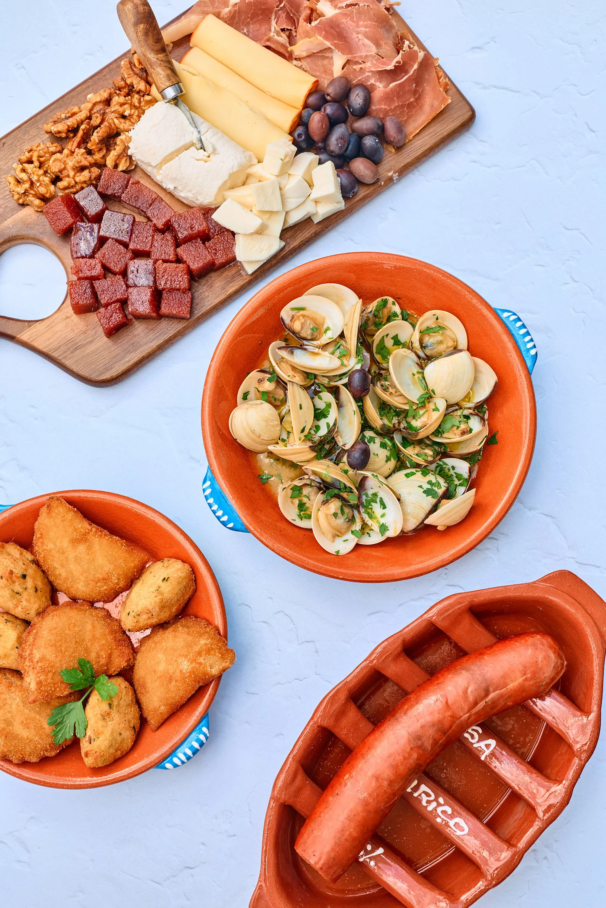 A wooden board with an assortment of cheeses, cured meats, black olives, and chopped cured meat. An orange bowl with clams garnished with parsley, a bowl of fried food, and a terracotta dish with sausages are also visible on a white surface.