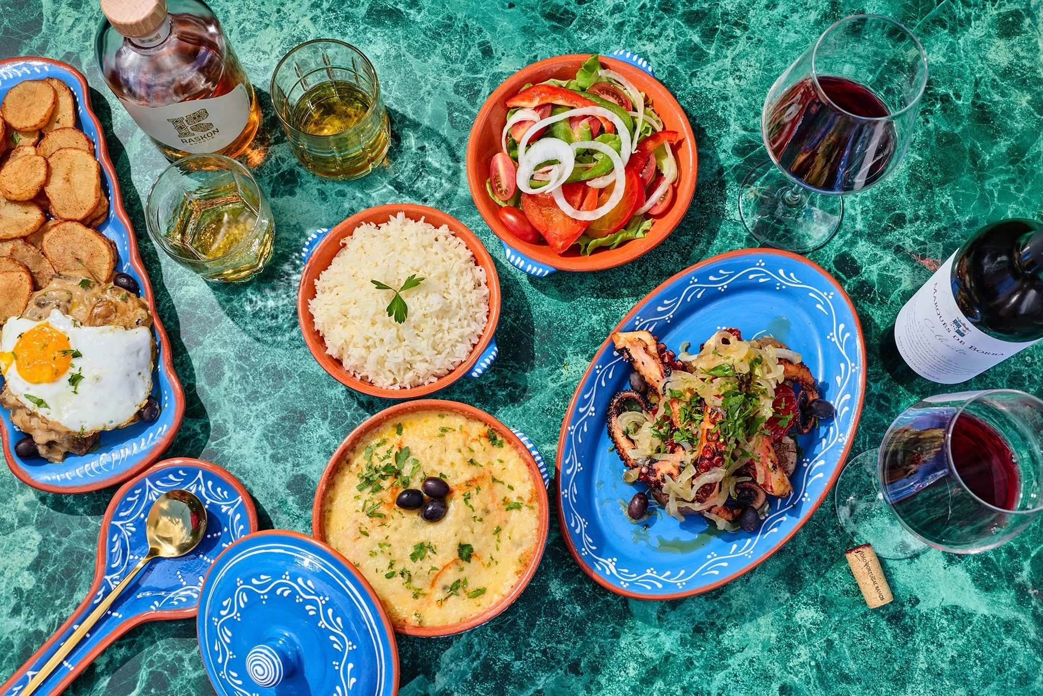 A colorful authentic meal on a green marble table, including a salad, rice, soup, and several side dishes with drinks of beer and red wine.