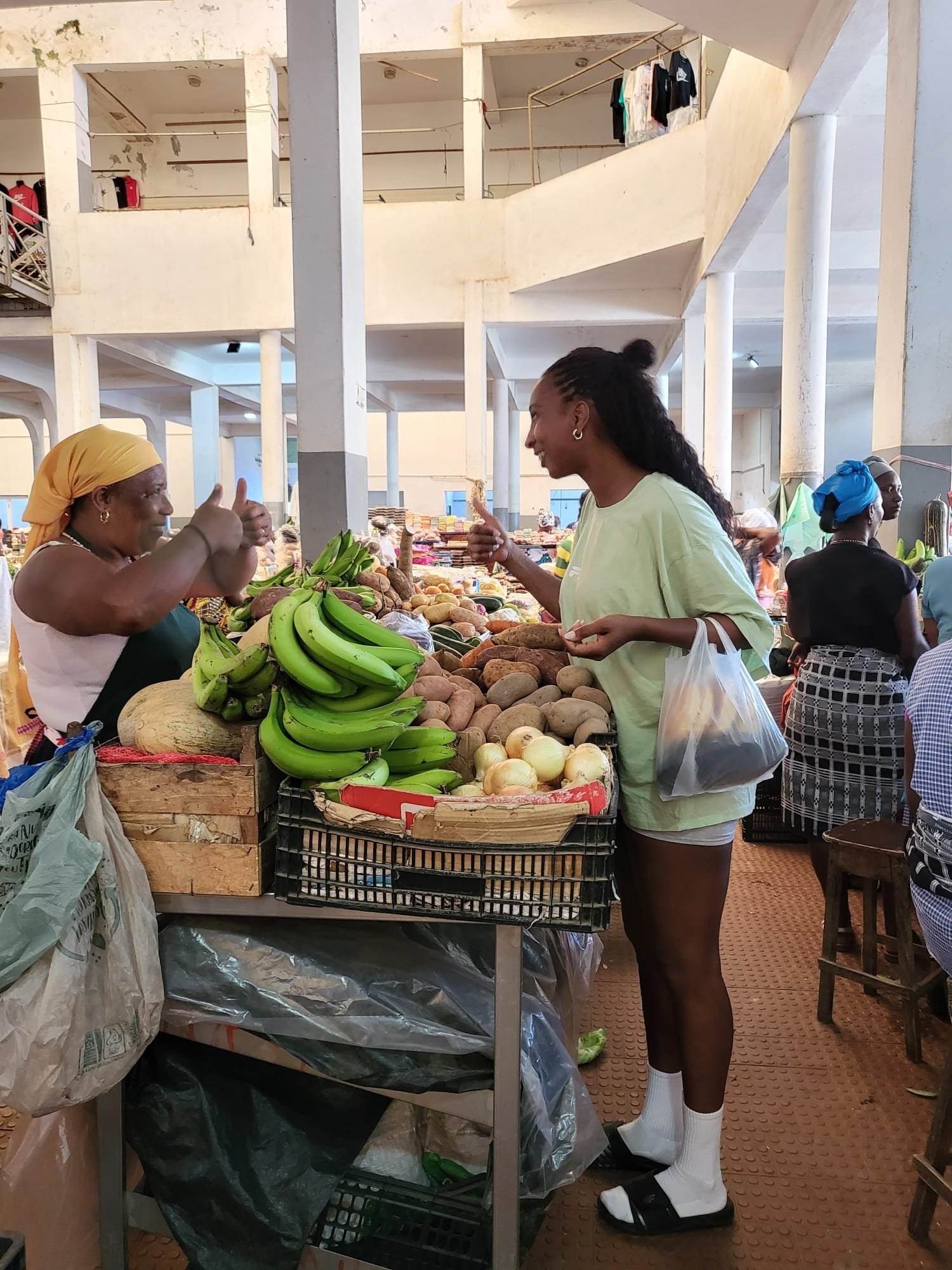 A young woman and an older woman are shopping at a produce market. The young woman is smiling and talking, while the older woman is giving a thumbs up. The market stall is filled with bananas, onions, and other vegetables. Other shoppers are visible 