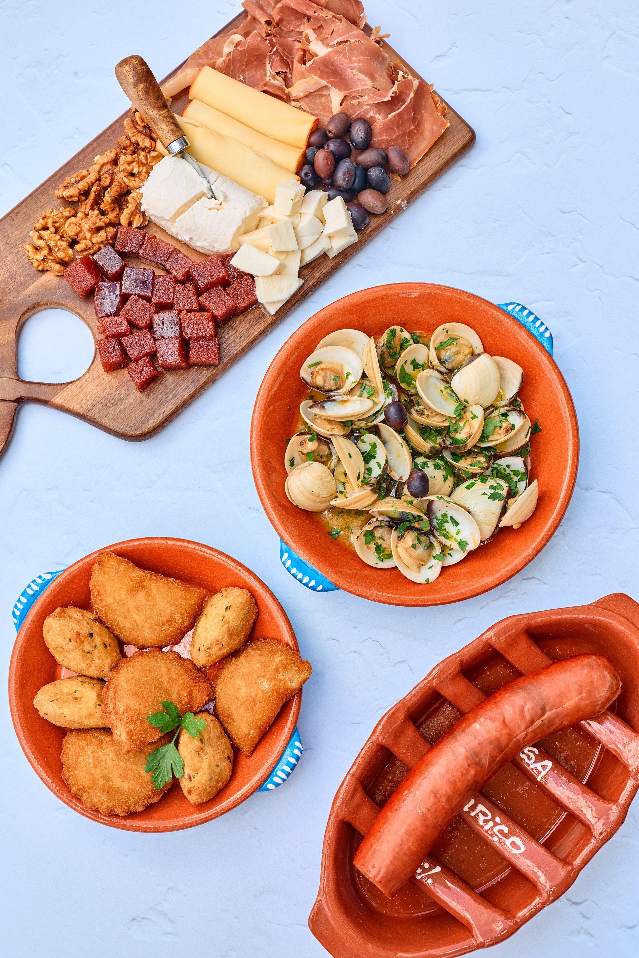 A charcuterie board with assorted cheeses, meats, grapes, walnuts, and a cheese knife, next to bowls of fried foods, clams, and hot dogs.