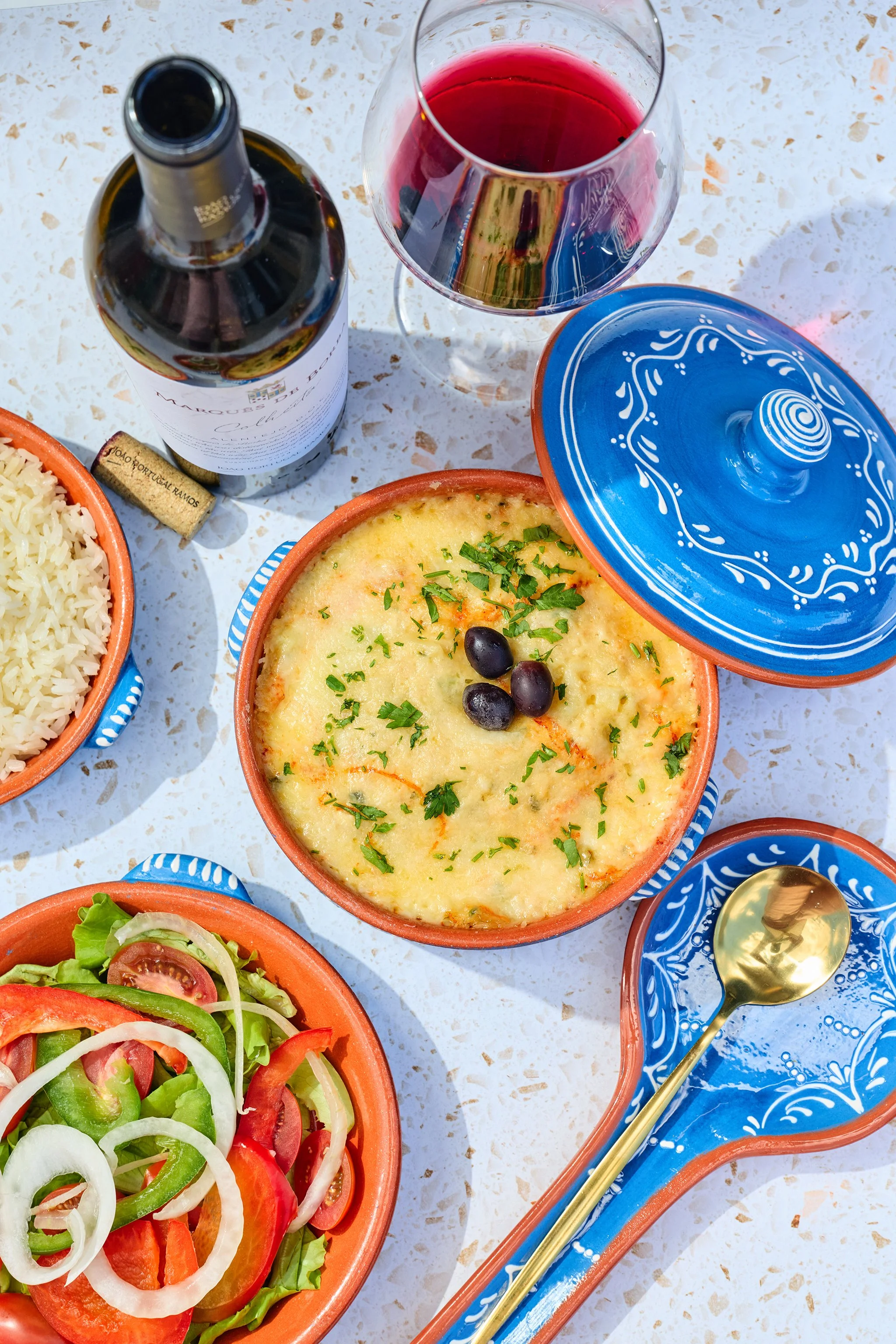 A table with a bowl of salad, a bowl of baked casserole topped with herbs and olives, a plate of mixed salad, a bottle of red wine, and a glass of red wine. Blue decorative dish covers and a gold spoon are also present.