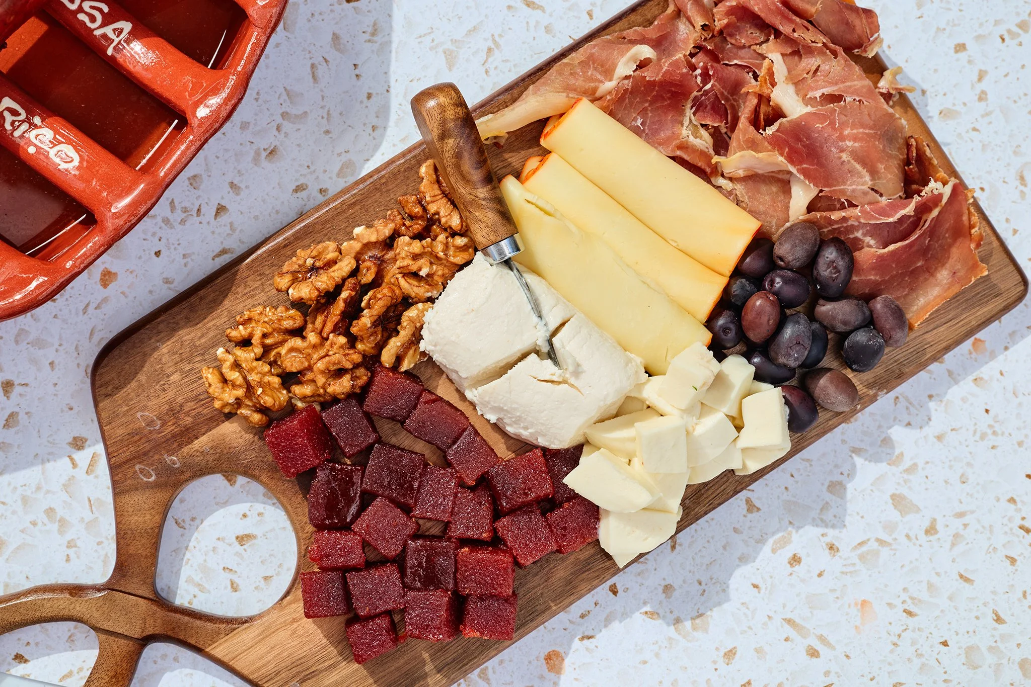 A wooden serving board with an assortment of cheeses, meats, grapes, and walnuts, placed on a speckled white countertop. Red bell pepper slices are visible in the bottom left corner.