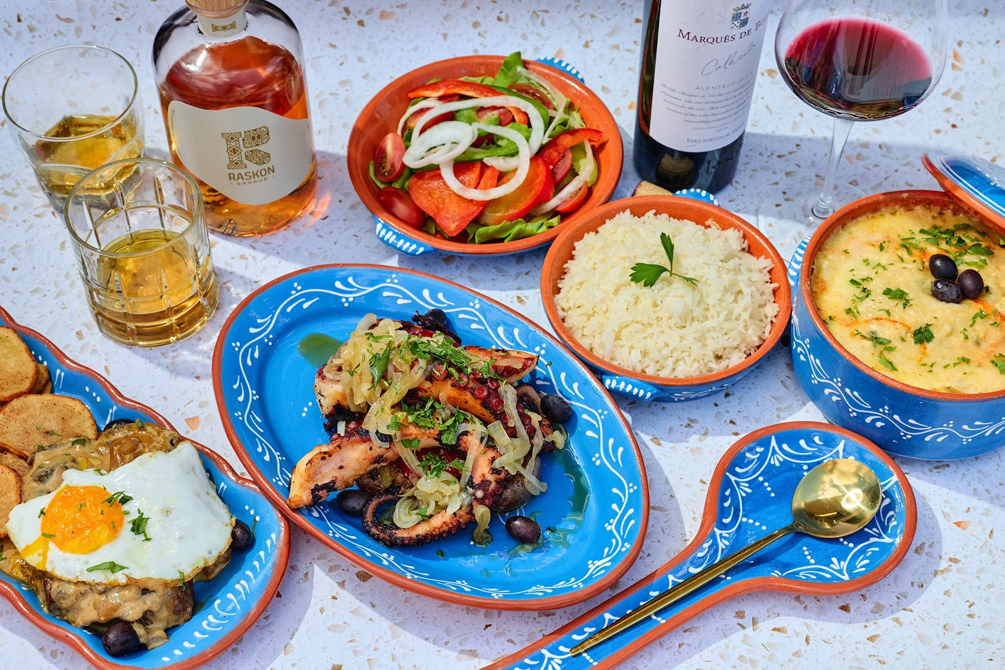 A colorful table setting featuring various dishes including a salad with tomatoes and onions, rice, a casserole topped with herbs and black olives, grilled meat with vegetables, a fried egg, and glasses of white and red wine, along with a bottle of wine and a bottle of liquor.