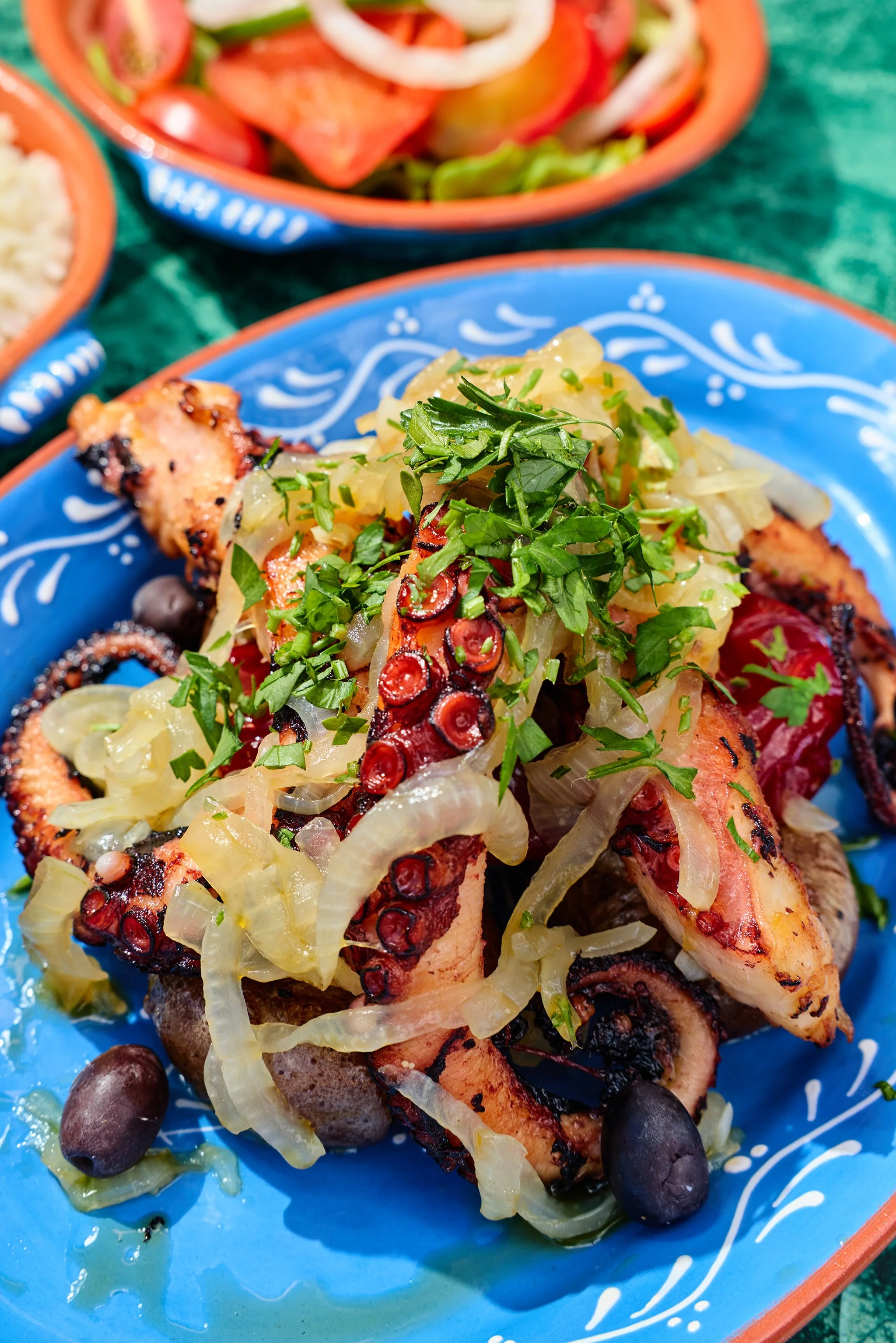 Grilled fish fillet topped with chopped herbs, sliced chili peppers, and pickled onions, served on a decorative blue plate with black olives and grilled vegetables in the background.