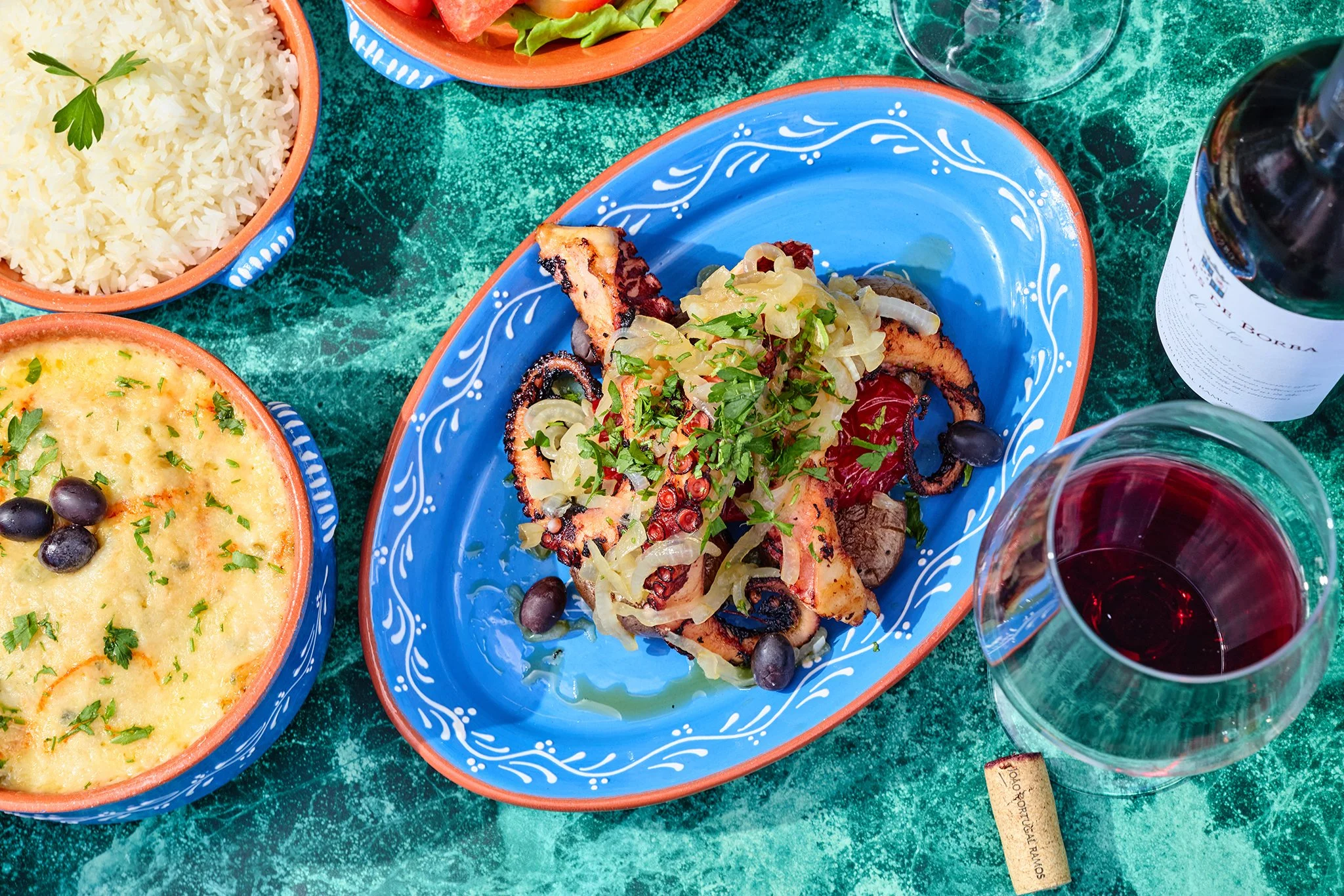 A table setting with colorful dishes of Mexican food, including rice, beans, grilled chicken topped with onions and cilantro, and shrimp, accompanied by glasses of red wine.