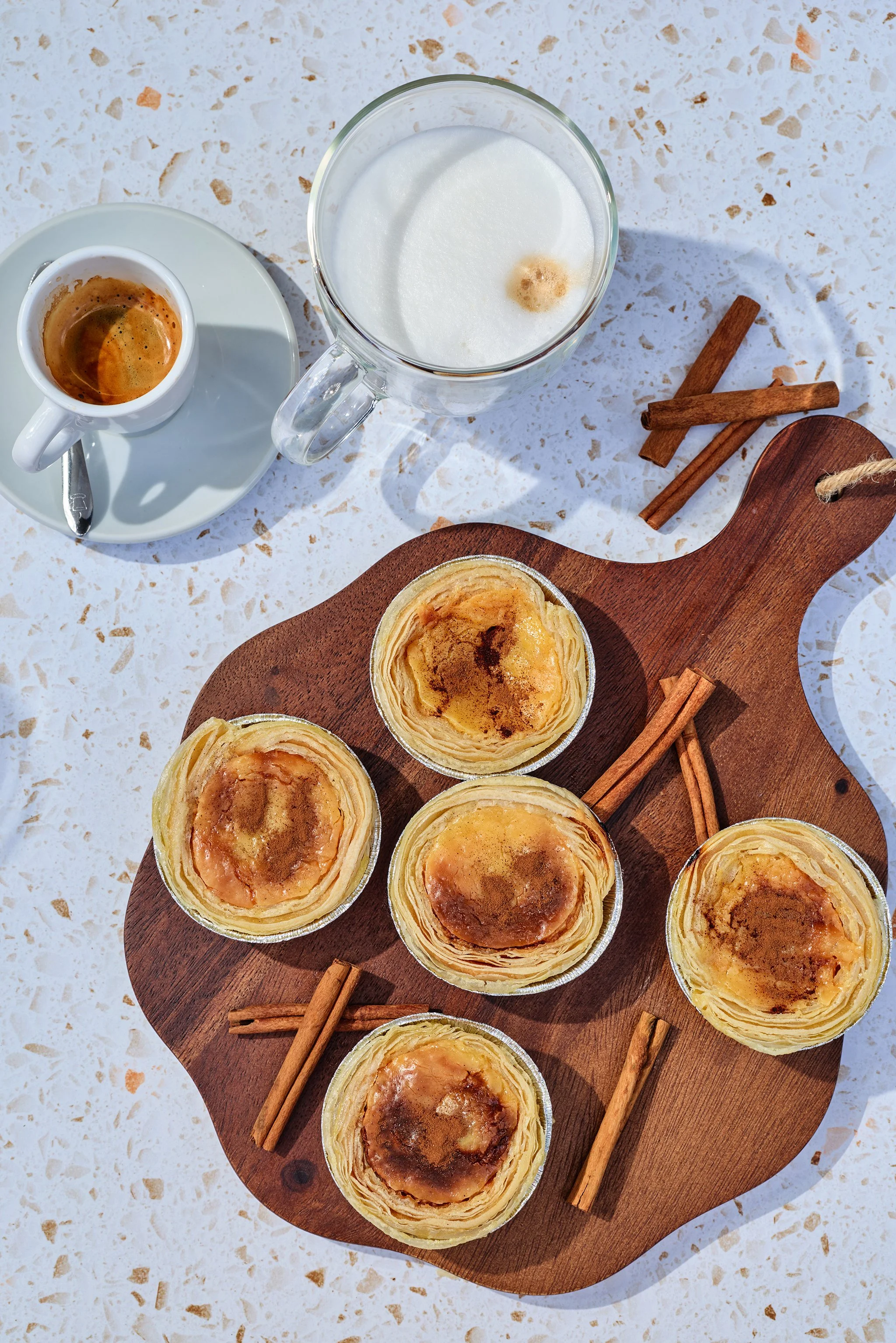 A top-down view of a wooden serving board with several baked egg tarts on it, on a speckled white surface. There is also a small cup of espresso, a glass of frothy milk, and two cinnamon sticks beside the board.