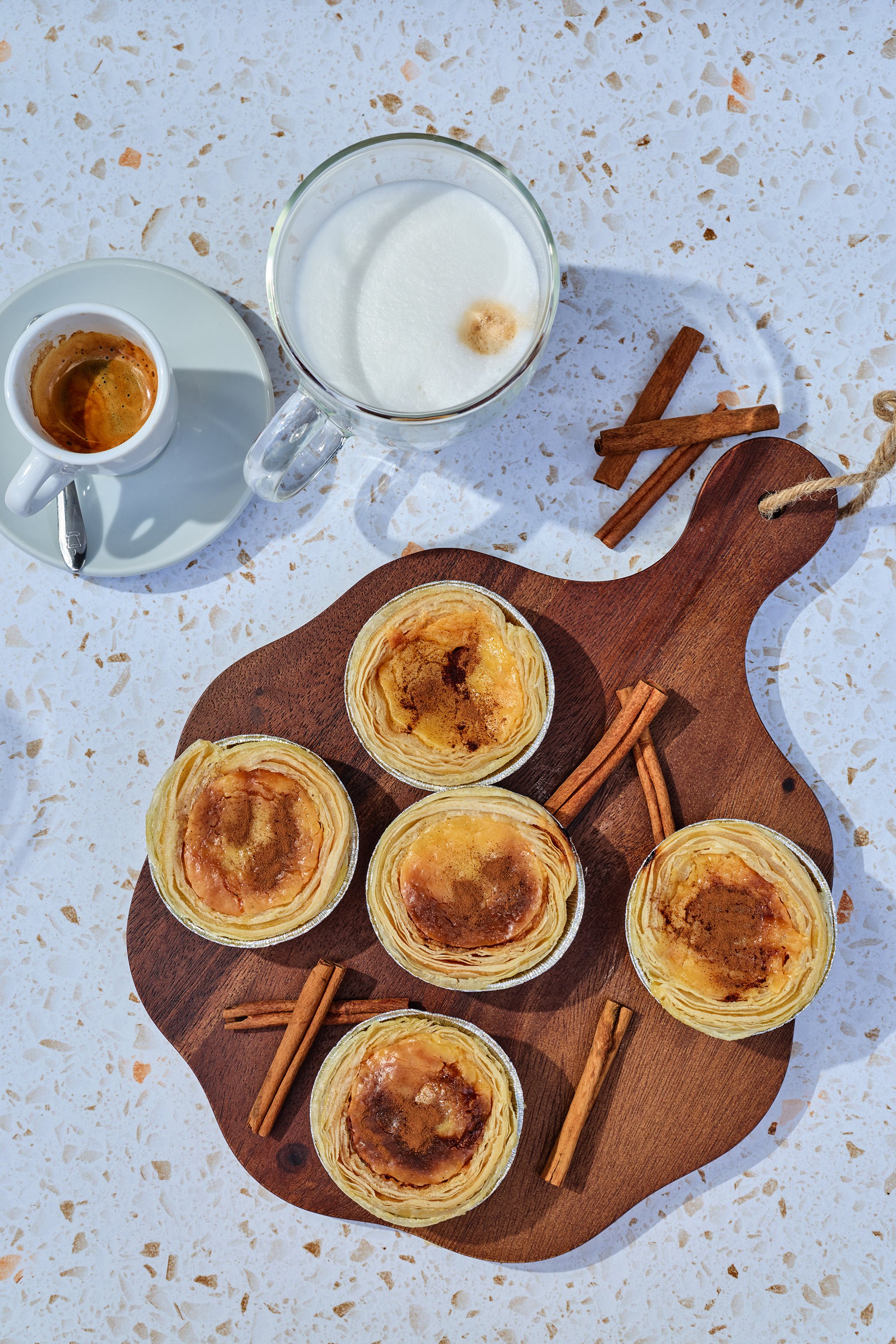 Top view of a wooden tray with Pastel de Nata, garnished with cinnamon sticks around the tray, a glass of milk with a small amount of foam, and a small cup of espresso on a white speckled surface.