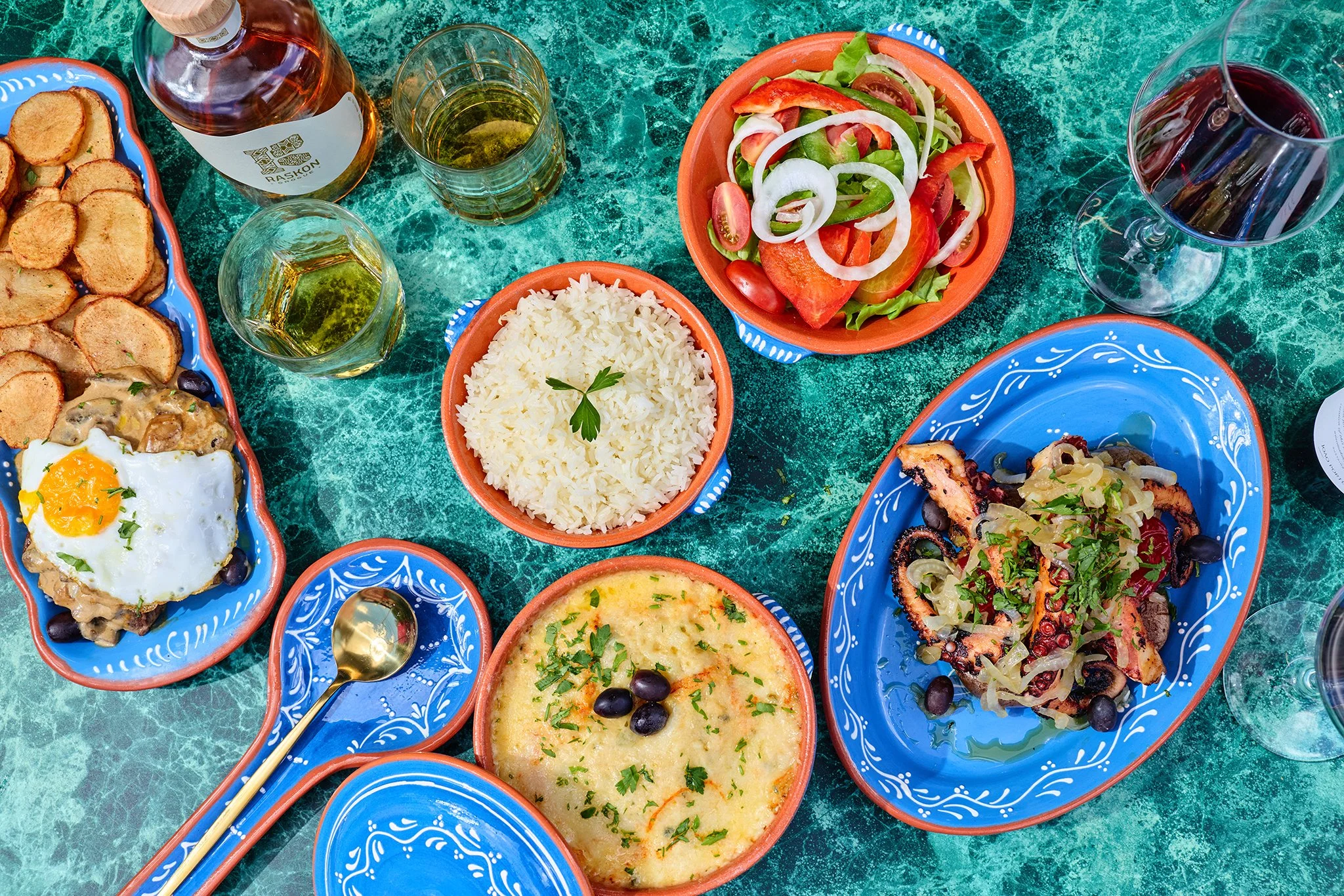 A colorful Portuguese meal spread on a green marble table includes rice, salad with tomatoes, onions, and lettuce, chicken with vegetables, two glasses of wine, a bottle of rosé, and a plate of fried plantains.