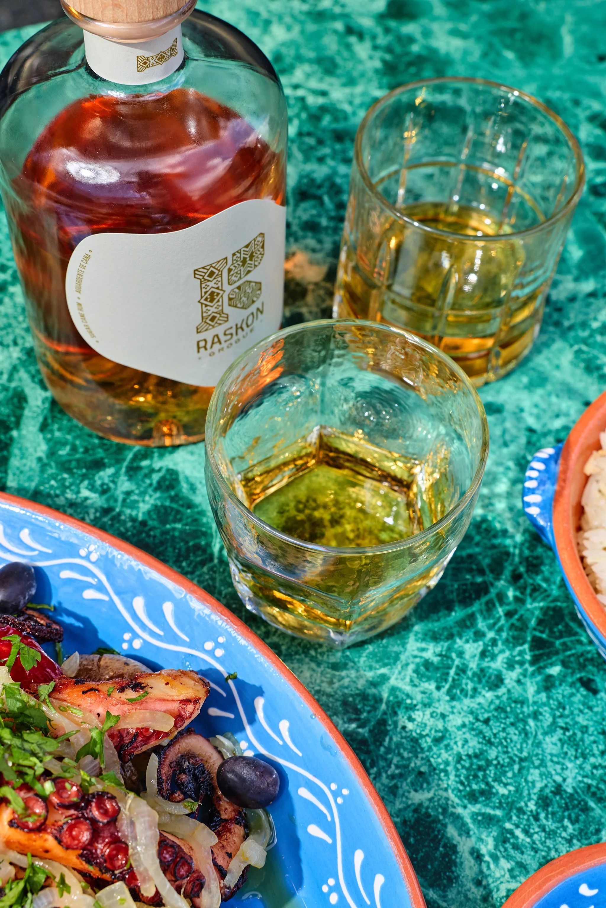 A bottle of RASKON brandy and two glasses with golden liquor on a green textured surface, along with part of a blue and orange patterned plate with food including octopus and vegetables.