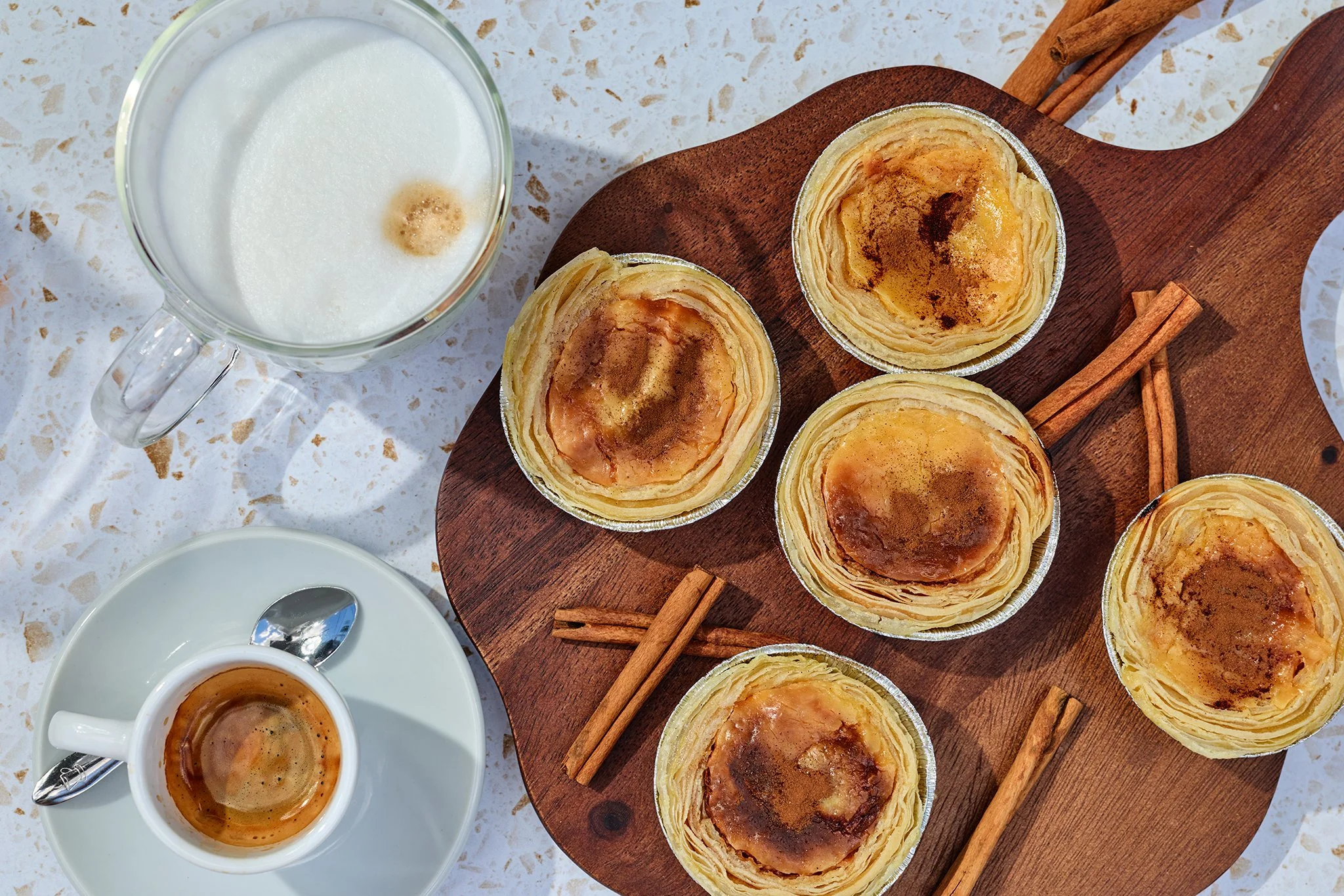 Pastries in a muffin tin on a wooden tray, with cinnamon sticks, a cup of coffee, a glass of milk, and a saucer with a spoon, on a white speckled surface.
