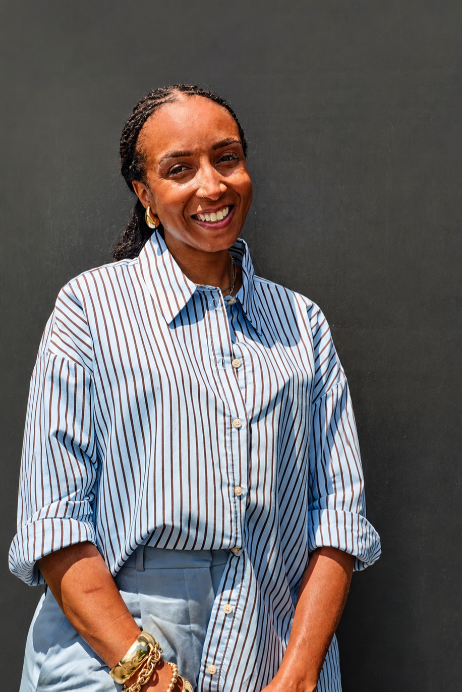 Smiling woman in a striped button-up shirt and gold jewelry standing against a dark wall.