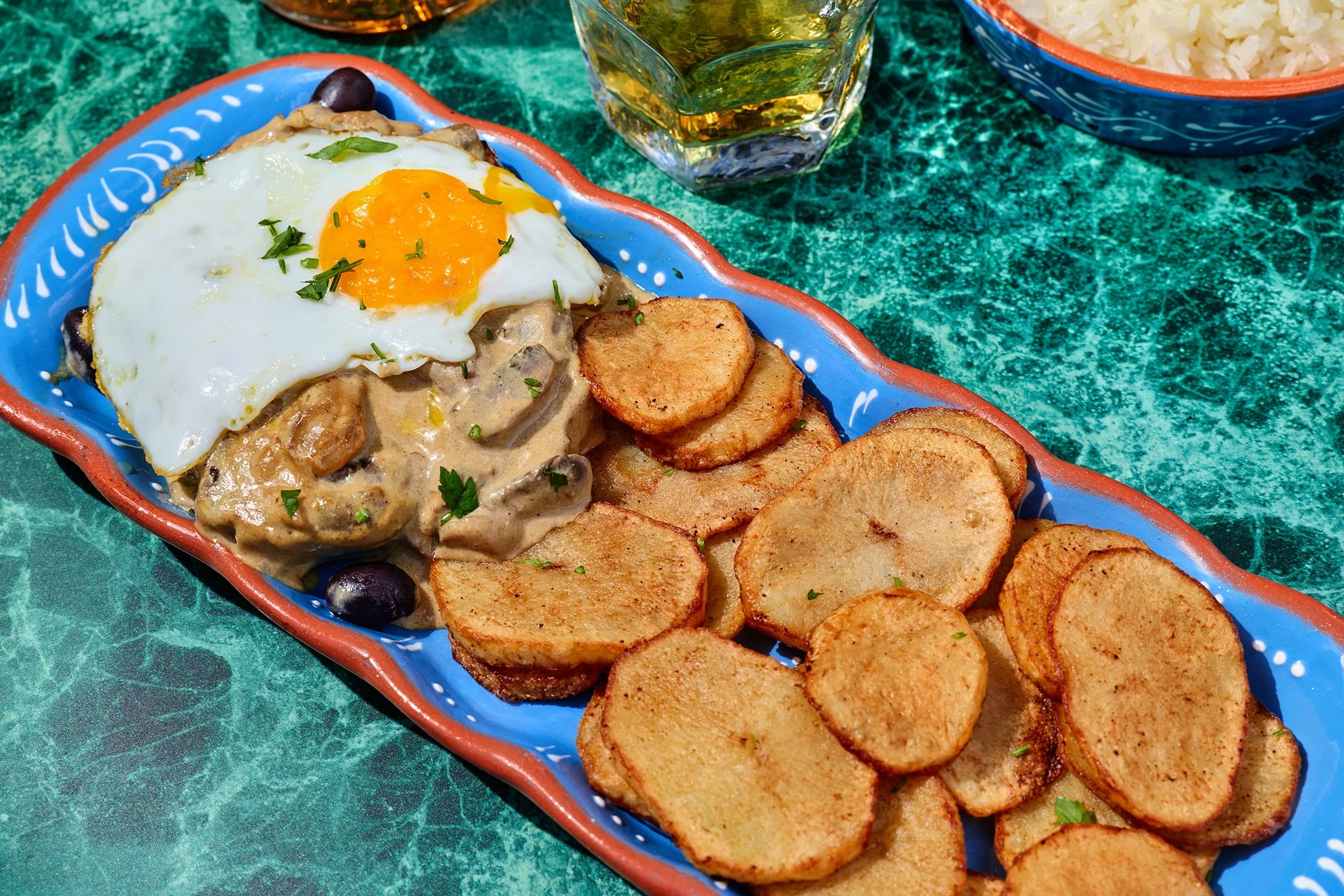 A colorful Mexican meal featuring a blue dish filled with fried plantains, a sunny-side-up egg topped with chopped herbs, and a creamy mushroom sauce with black beans, accompanied by a bowl of rice, a glass of beer, and a small bowl of salsa.