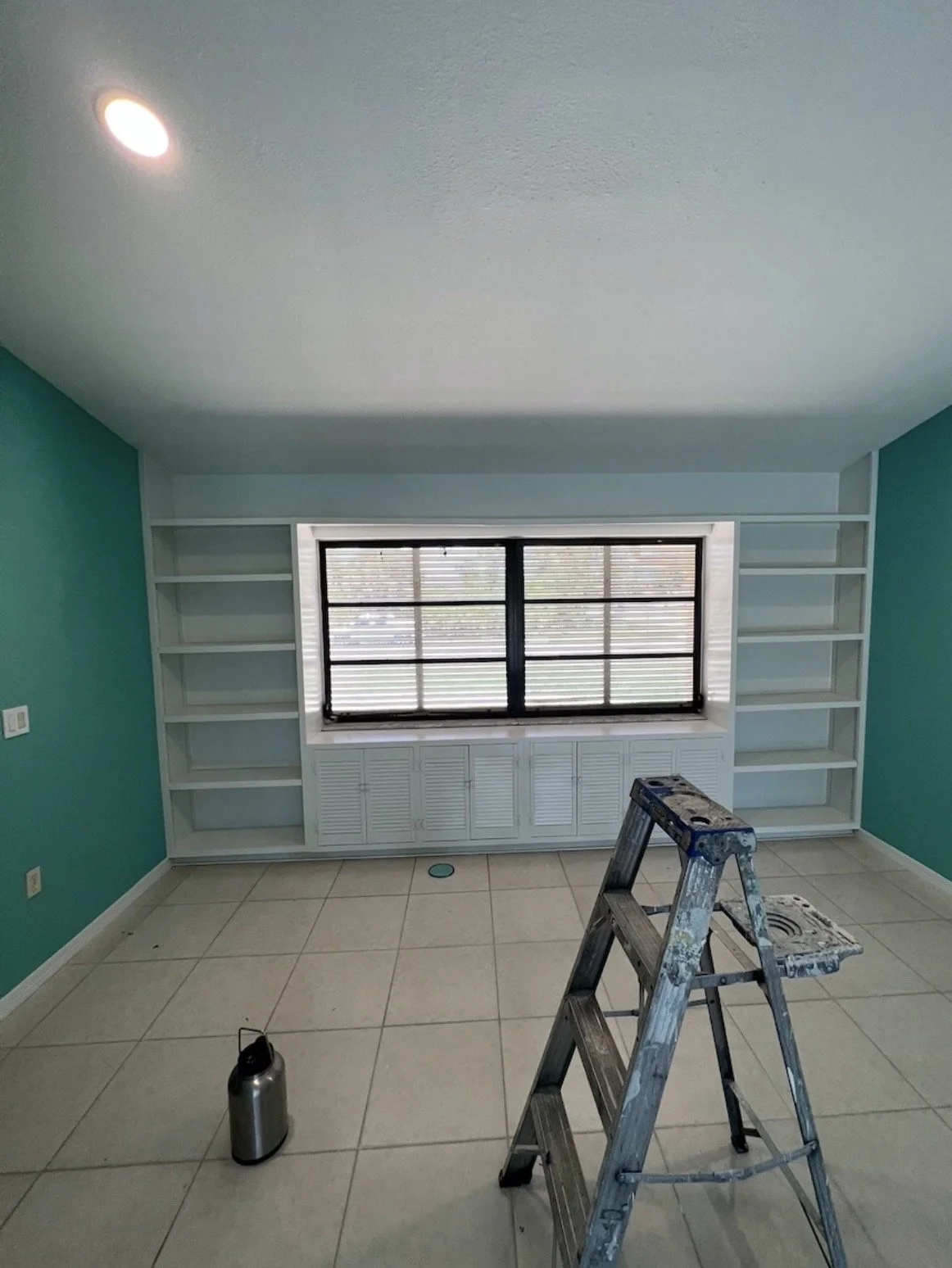 A room under renovation with turquoise walls, white built-in bookshelves, a large window with blinds, a step ladder, and a water bottle on the tiled floor.