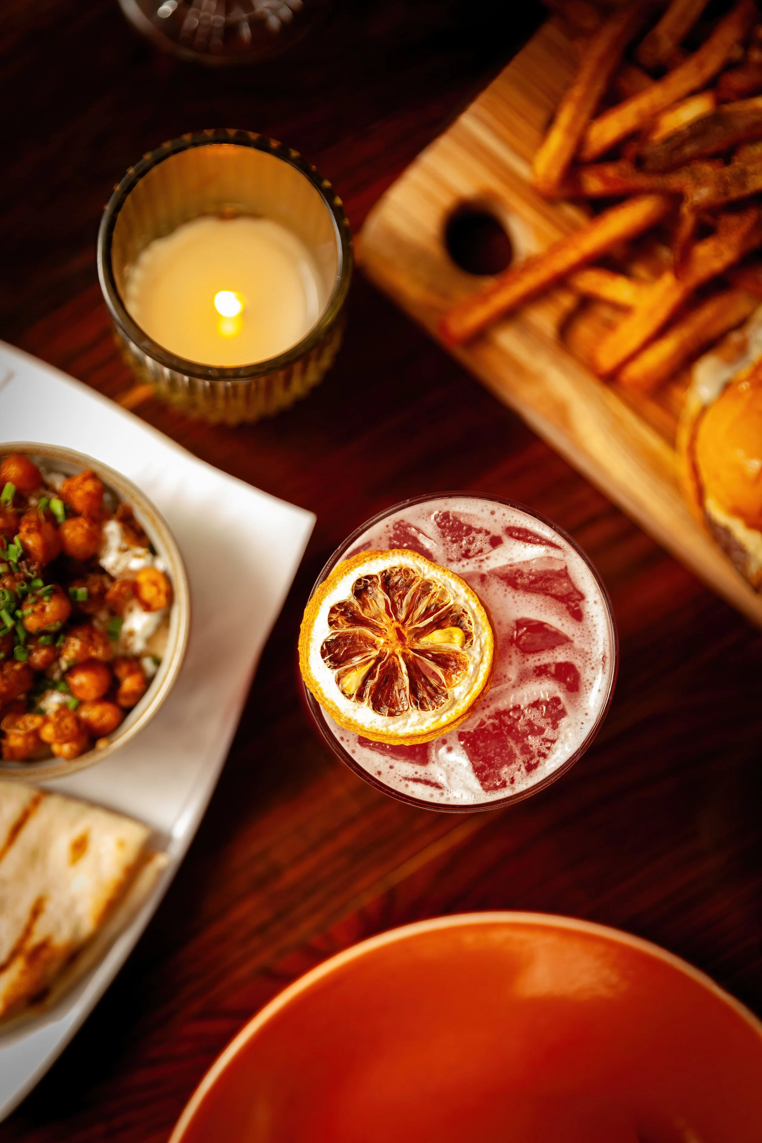 A top-down view of a table with a lit candle, a drink garnished with dried orange slices, a wooden tray with French fries, and a bowl of chickpea salad with herbs, all on a dark wooden surface.