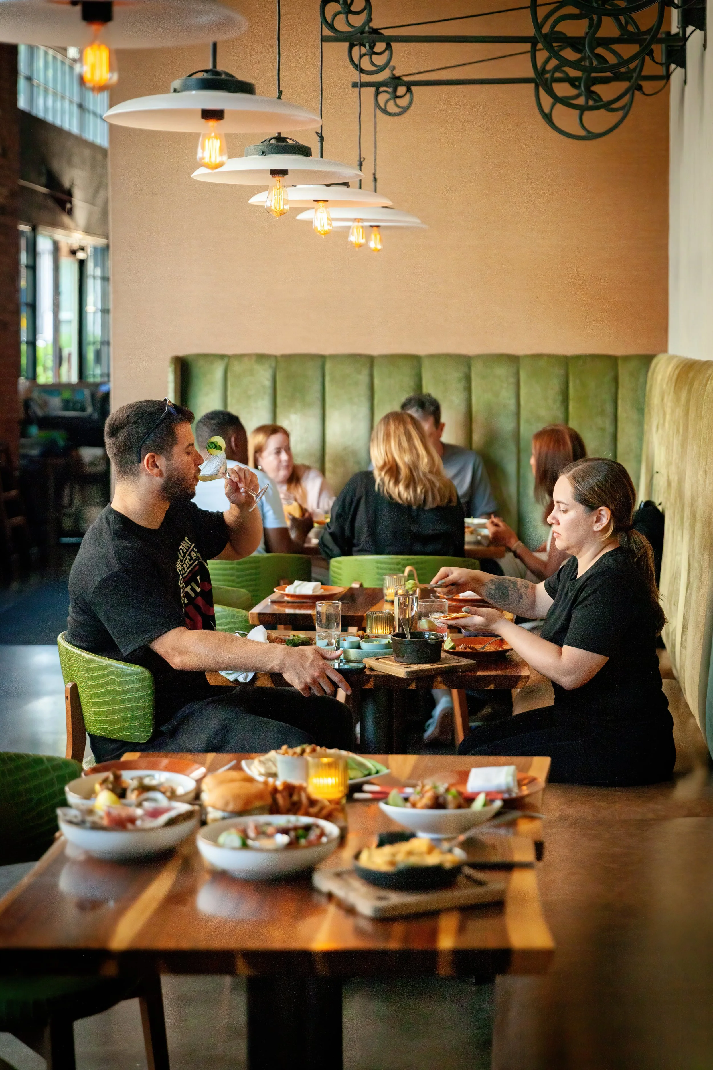 People dining at a restaurant with dishes of food on the tables and modern hanging lights above.