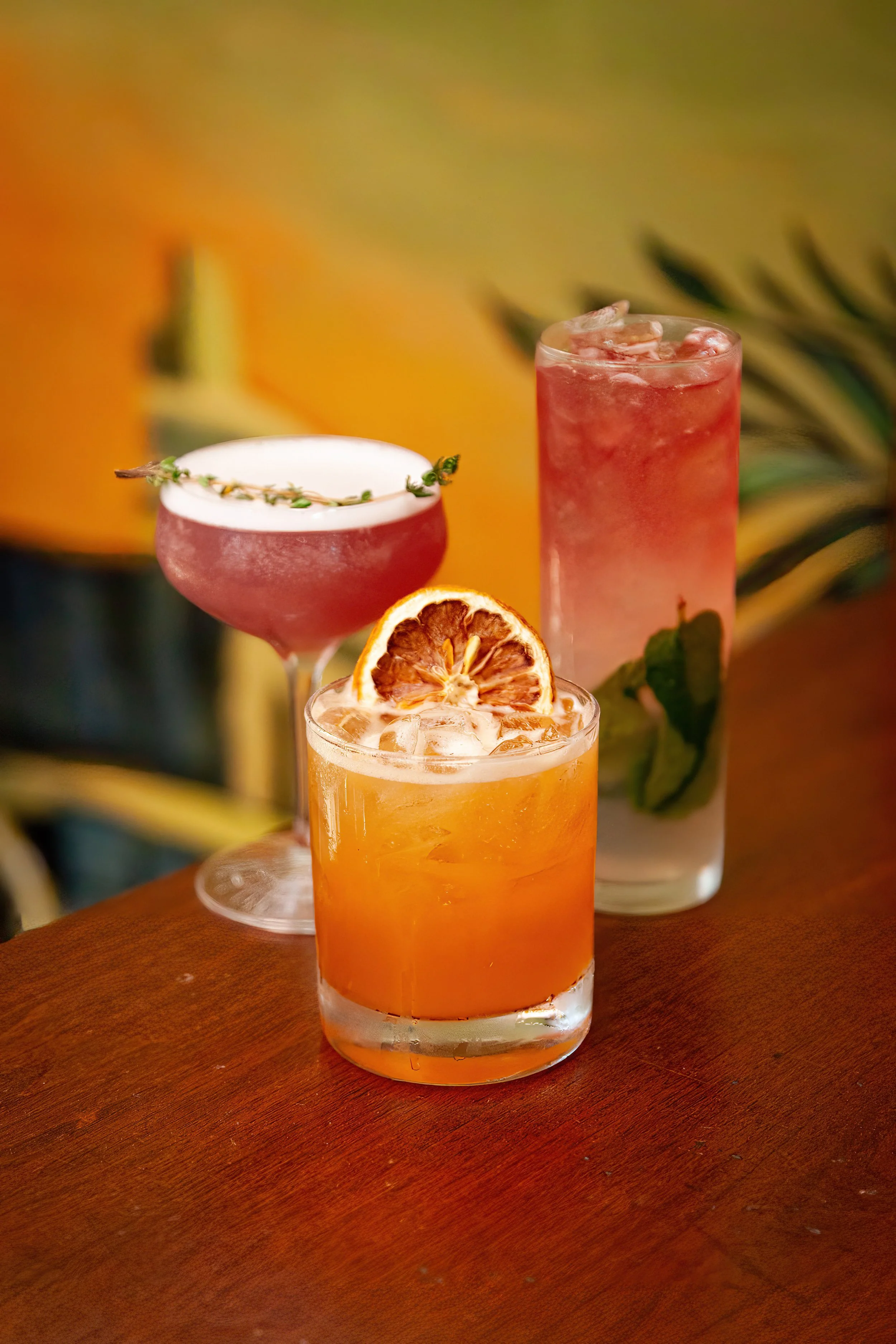 Three colorful cocktails on a wooden table with tropical background.