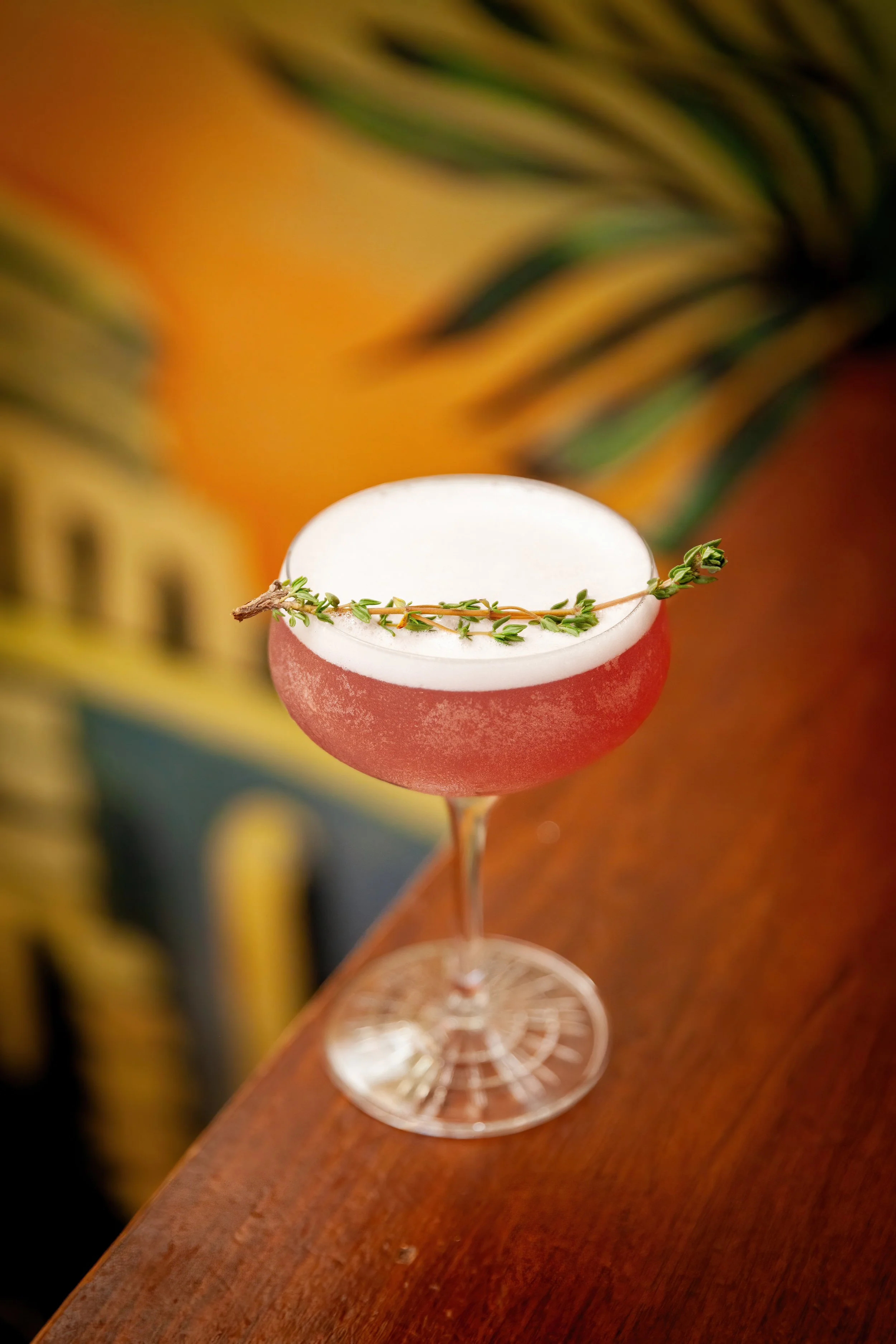 A cocktail in a coupe glass garnished with a sprig of thyme, with a blurred colorful tropical background.