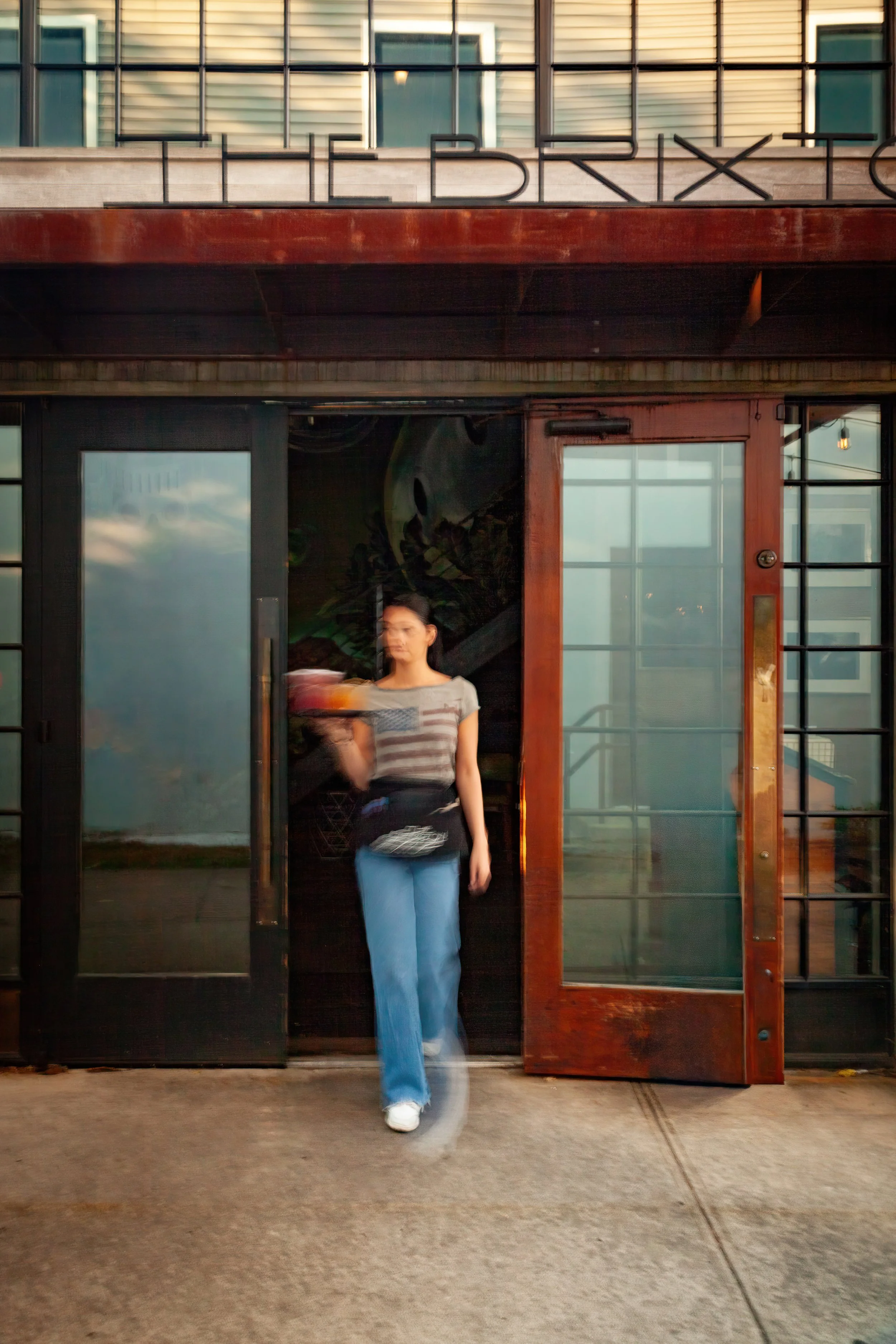 Person walking out of a restaurant or cafe with a glass door, carrying a drink, wearing a striped shirt and blue jeans.