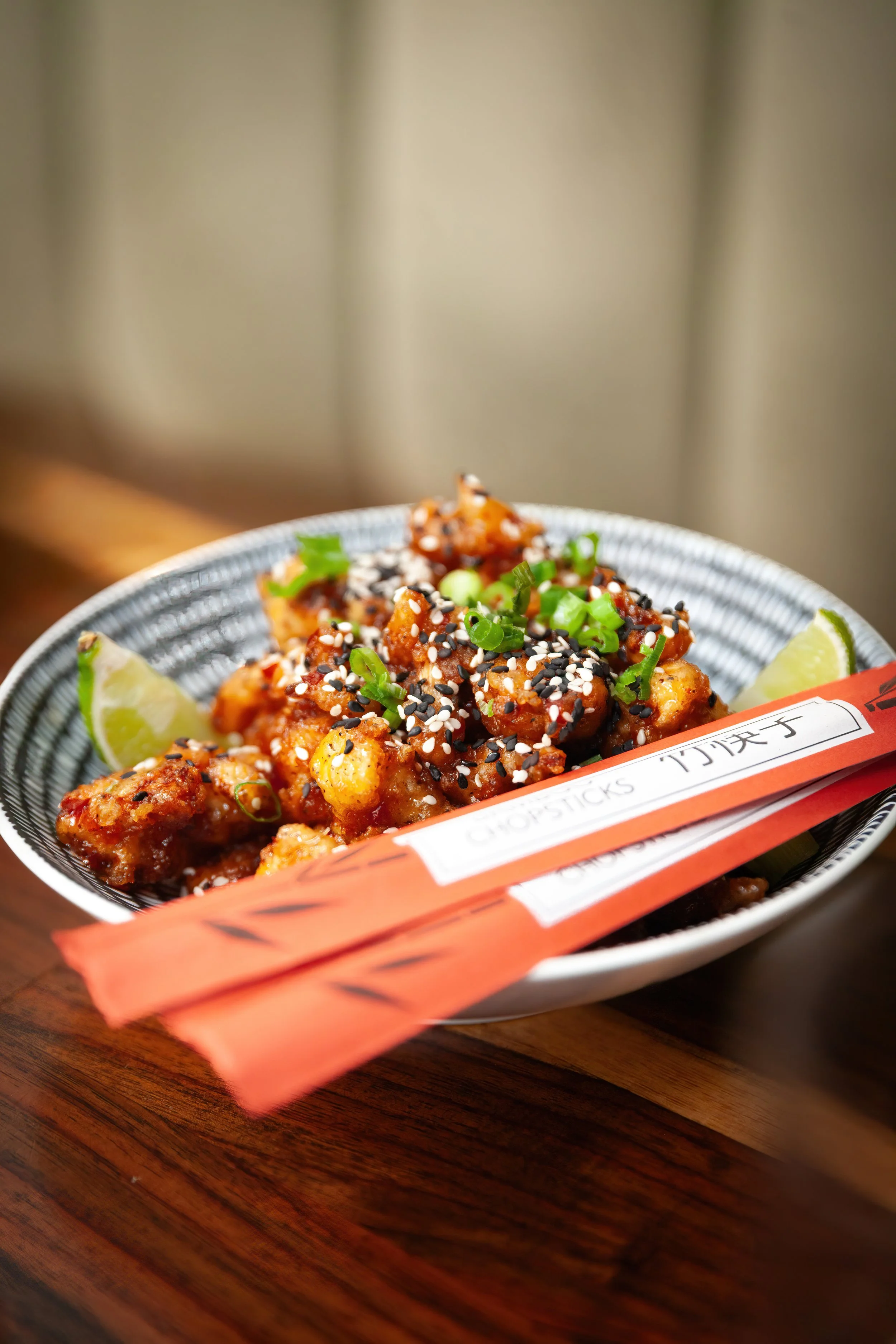 Close-up of a bowl of sushi topped with black and white sesame seeds, chopped green onions, and lemon wedges, with orange chopstick packaging in the foreground.