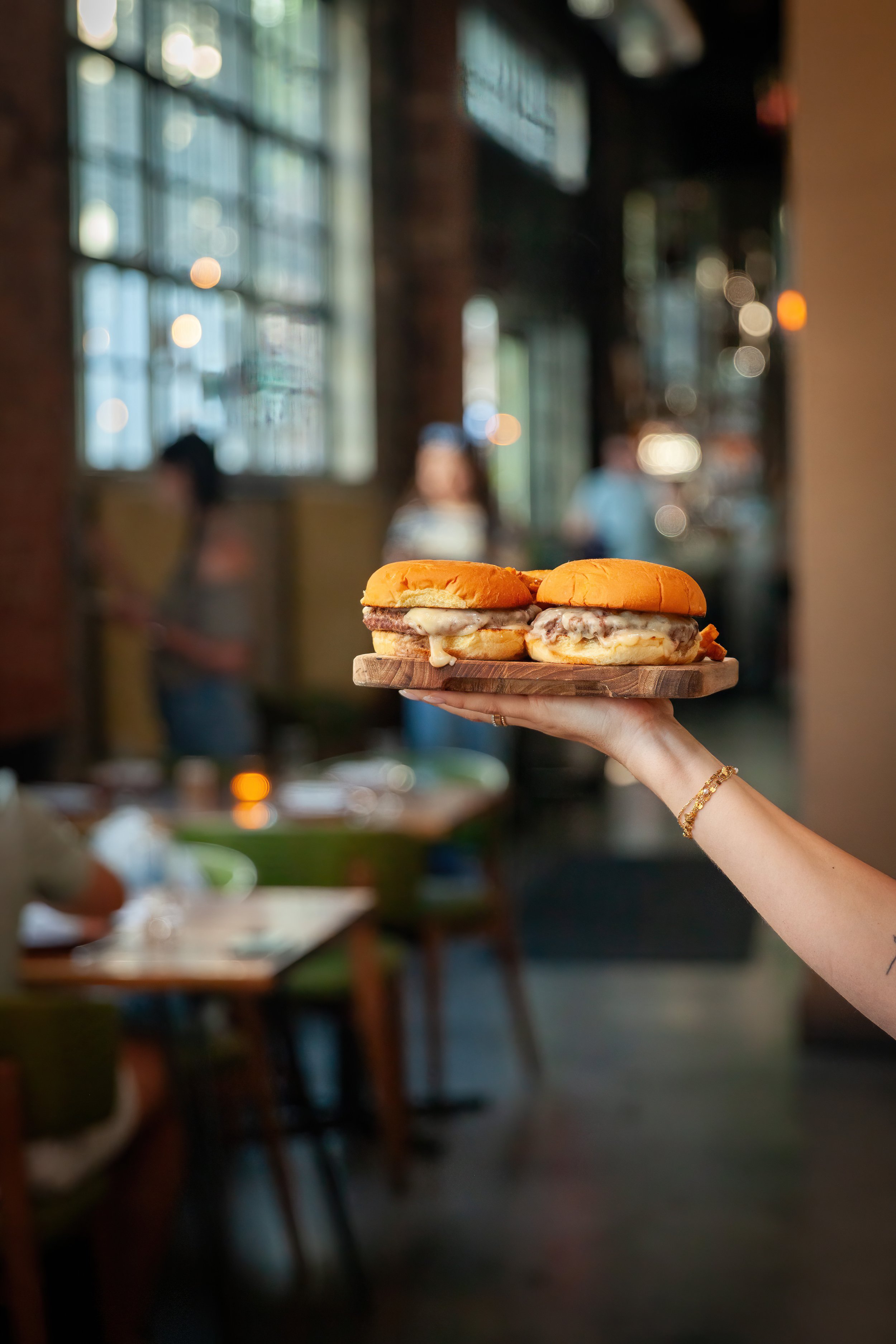 A hand holding a wooden tray with two cheeseburgers in a restaurant with a blurred background of large windows and tables.