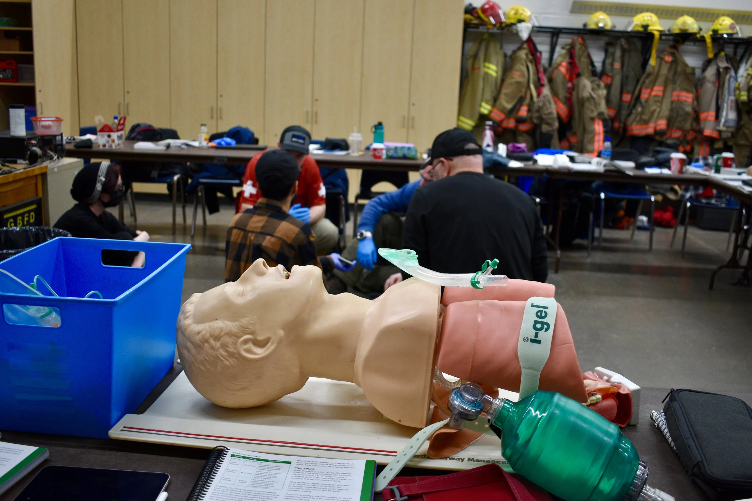 Firefighter medical training session with a CPR mannequin, intubation equipment, and protective gear in the background