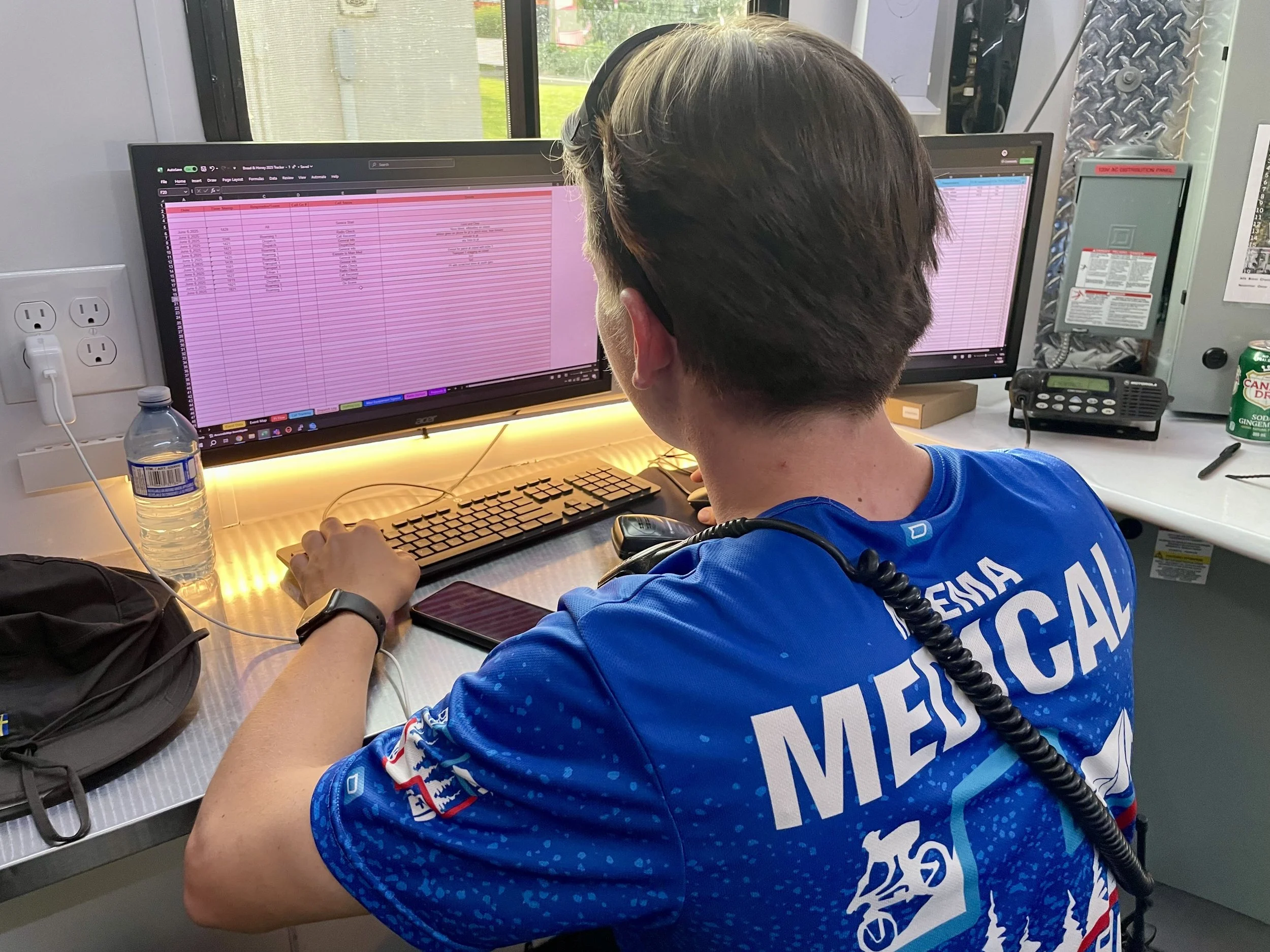 NPEMA dispatcher in a blue uniform working at a computer workstation with dual monitors, keyboard, and radio.