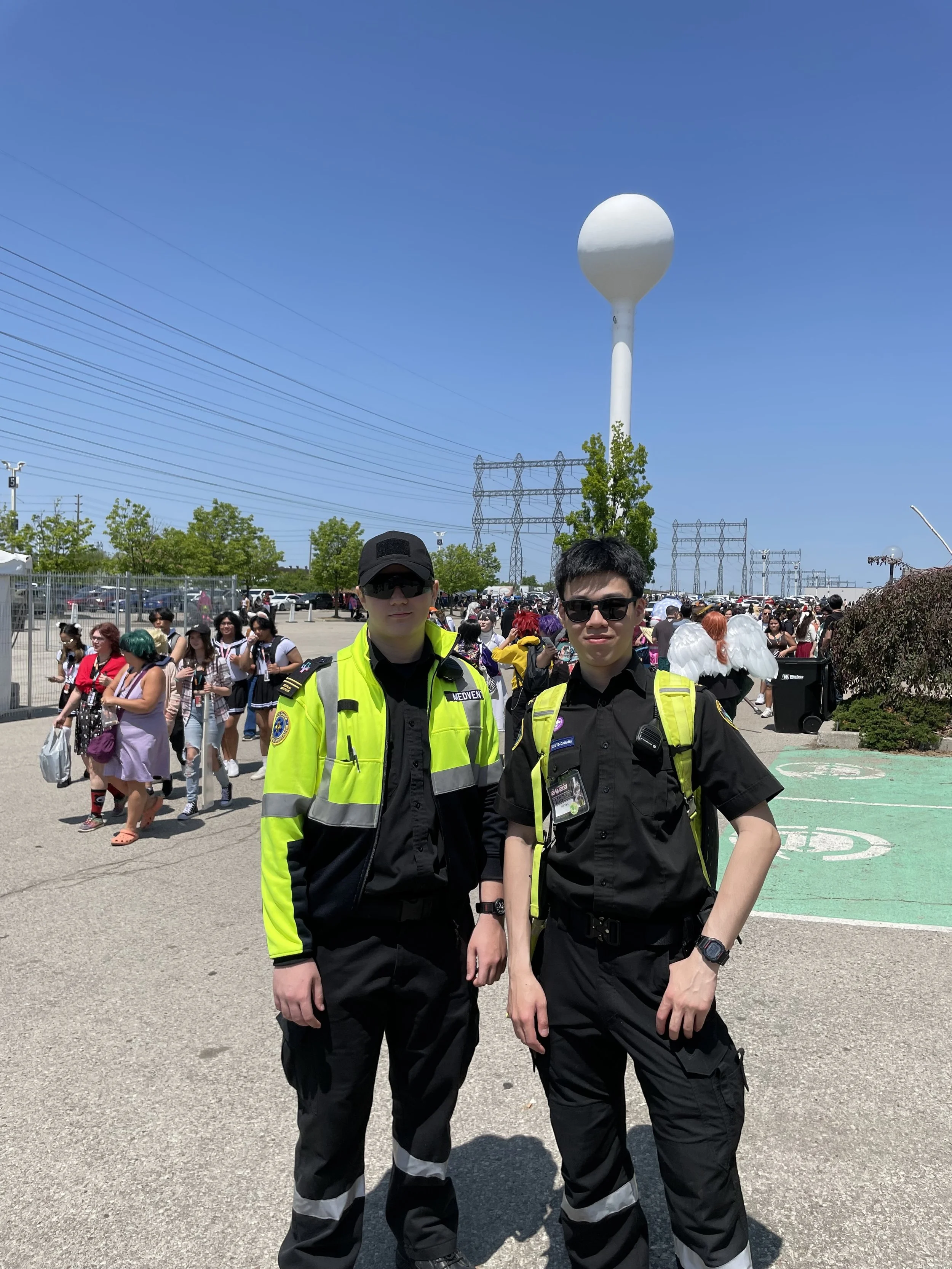 Two event medics in high-visibility uniforms pose at an outdoor festival beside a crowd, utility towers, and a large white water tower.