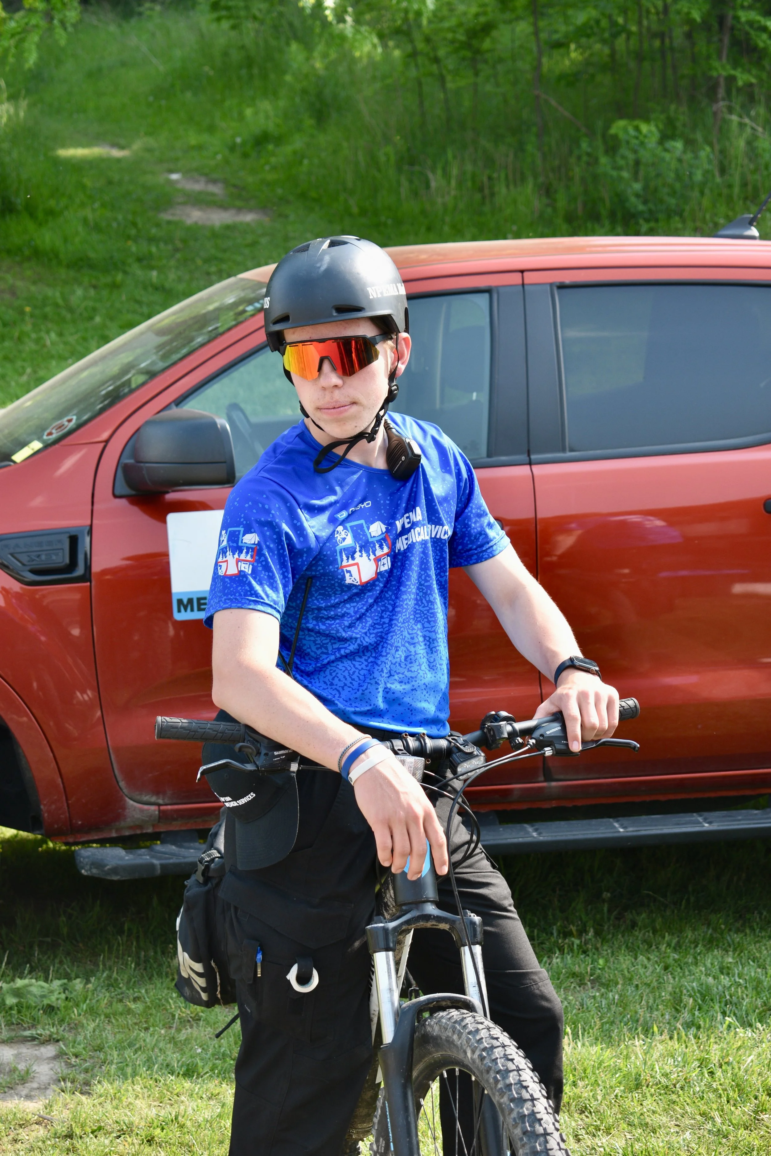 NPEMA mobile bike team responder  wearing a helmet and orange sunglasses sits on a mountain bike in front of a red vehicle in a wooded area.