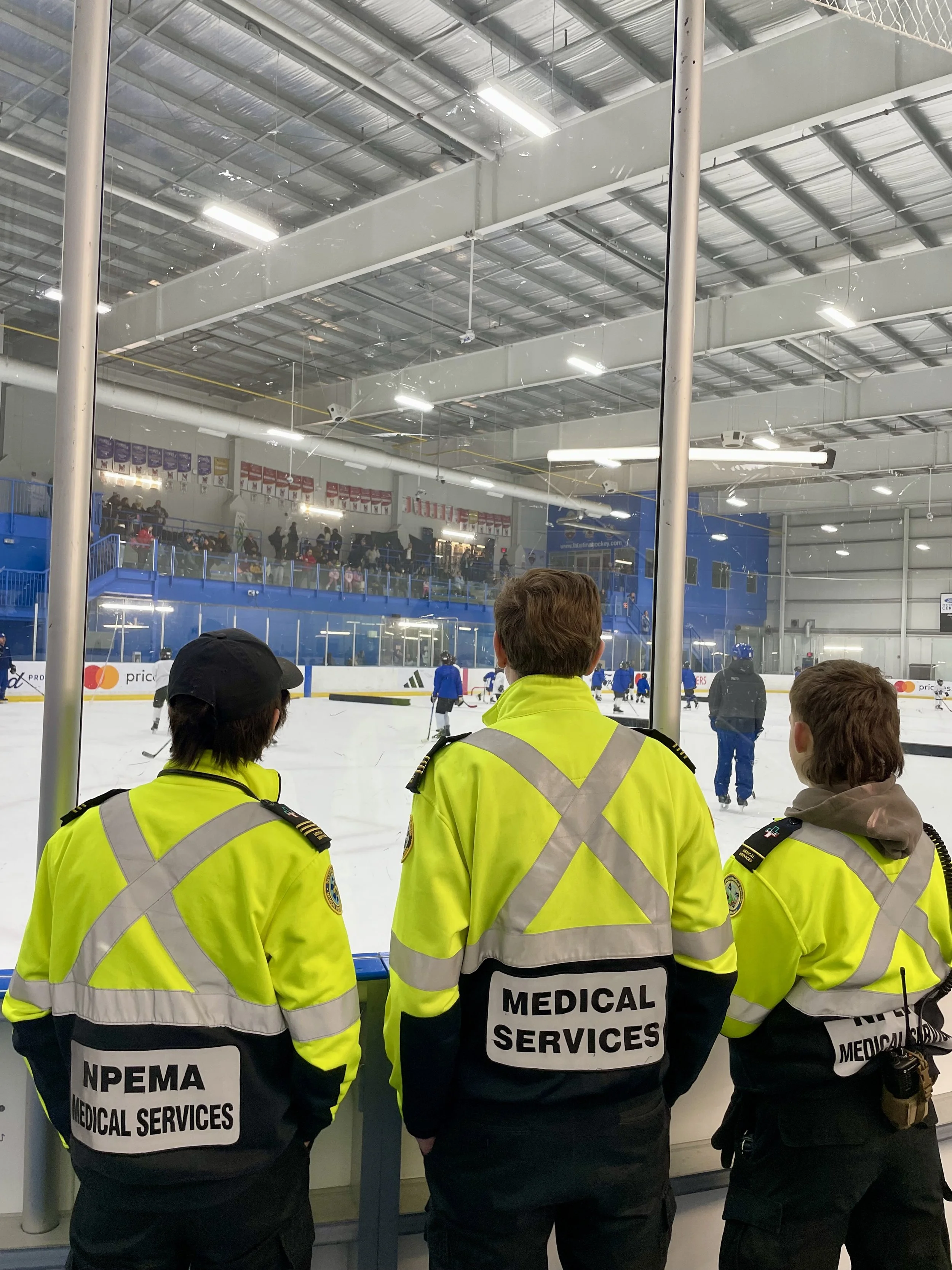 Medical services staff in high-visibility jackets watch a youth hockey practice from behind the rink glass.