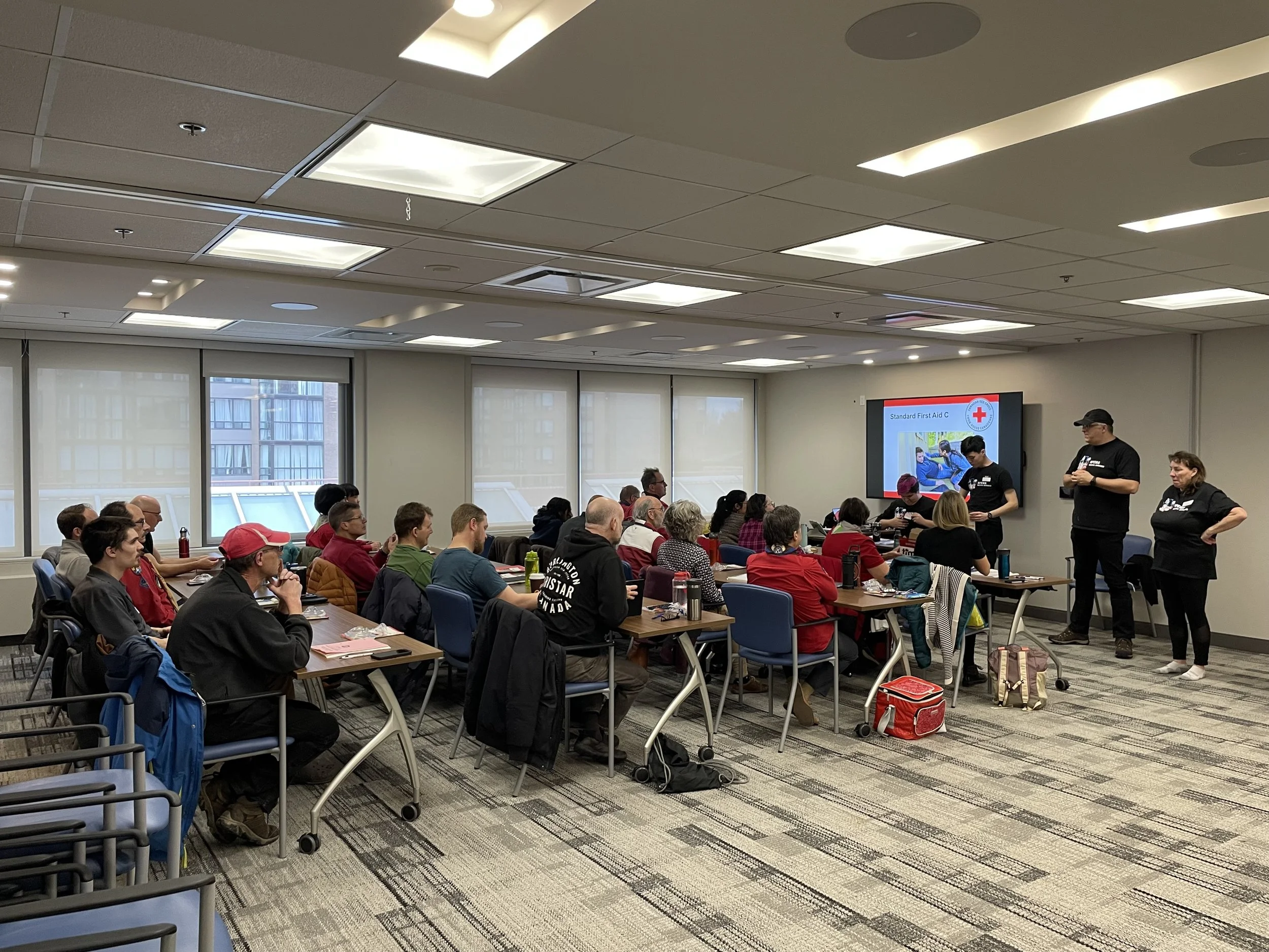 First aid training class with seated participants watching instructors and a Standard First Aid presentation in a conference room