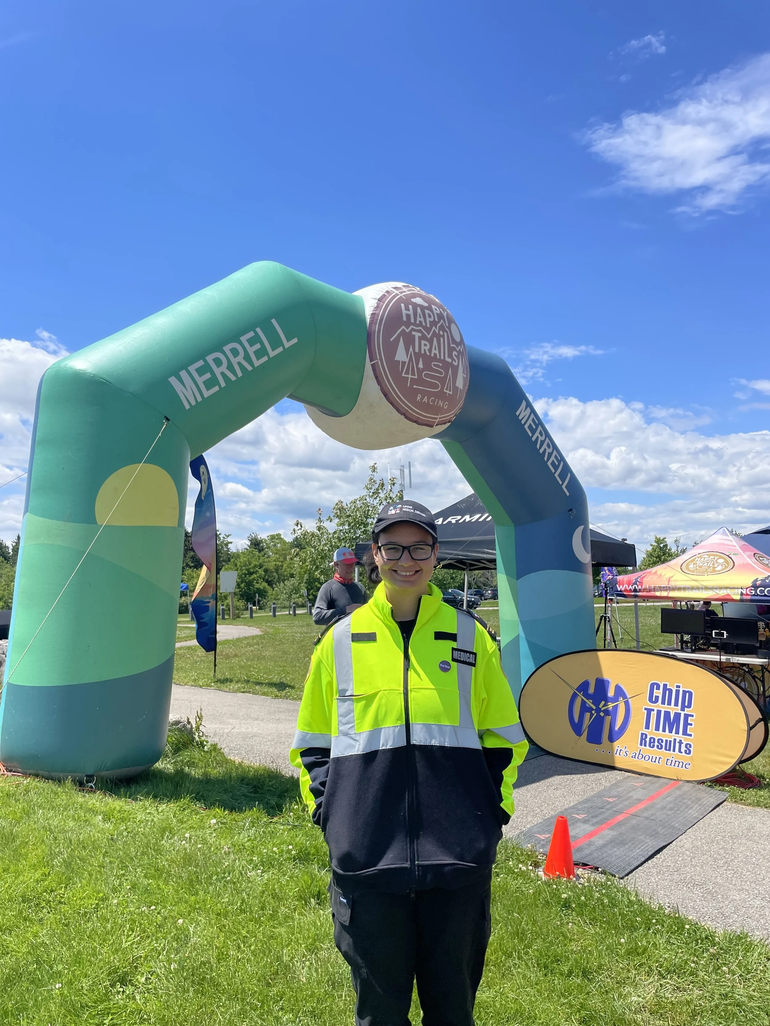 Smiling npema medical responder volunteer in a high-visibility jacket standing beneath a Happy Trails race arch at an outdoor running event.